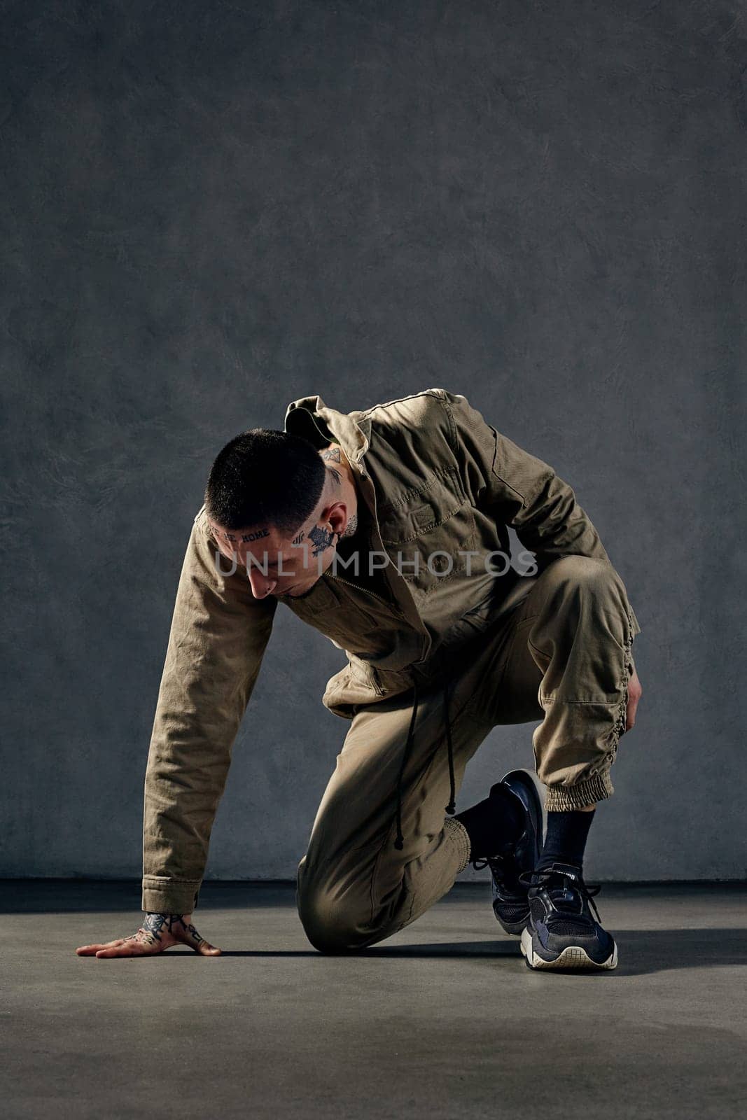 Unusual male with tattooed body and face, earrings, beard. Dressed in khaki jumpsuit, black sneakers. Dancing on gray background. Dancehall, hip-hop by nazarovsergey