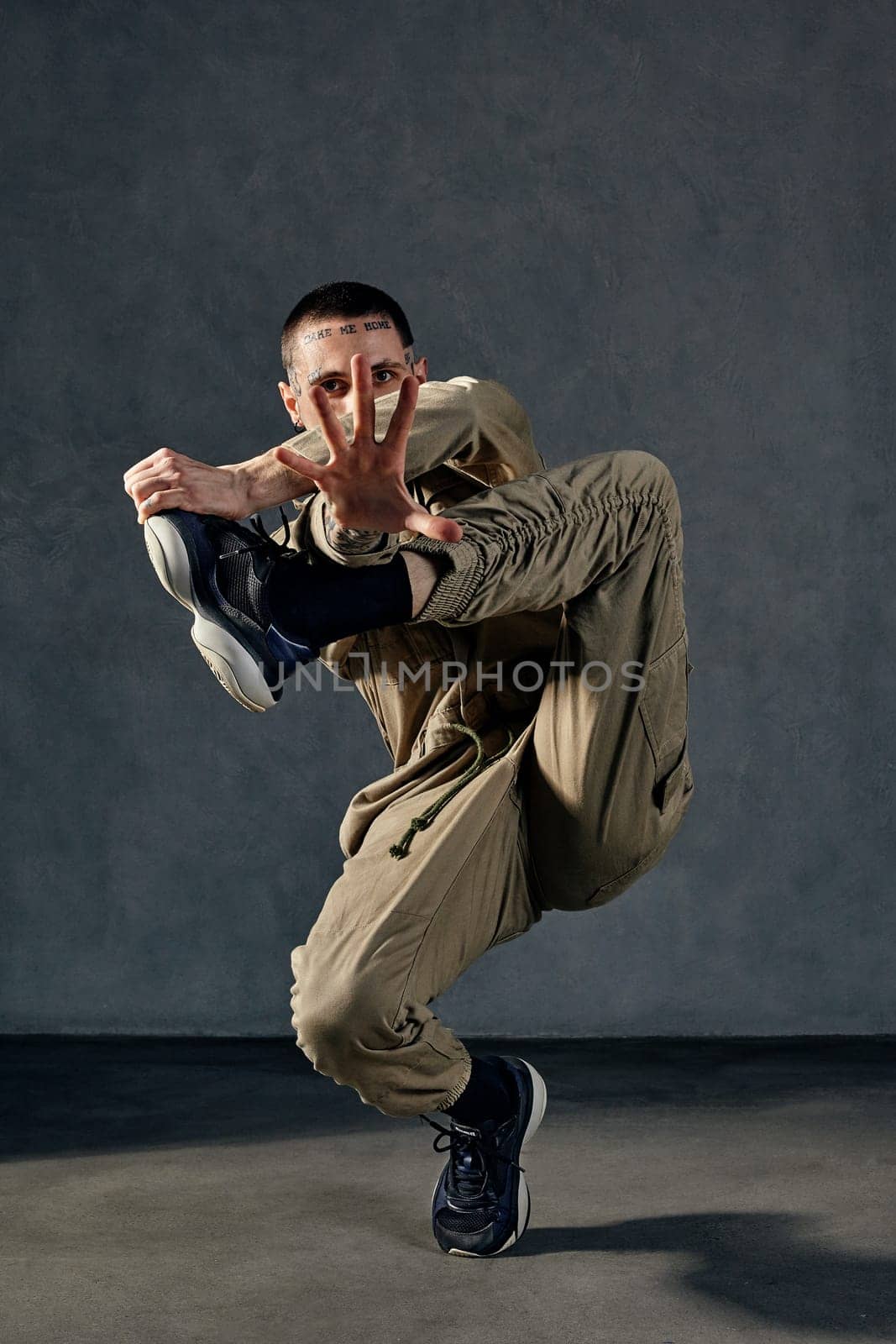 Young unusual guy with tattooed body and face, earrings, beard. Dressed in khaki overalls and black sneakers. He is dancing against gray studio background. Dancehall, hip-hop. Full length, copy space