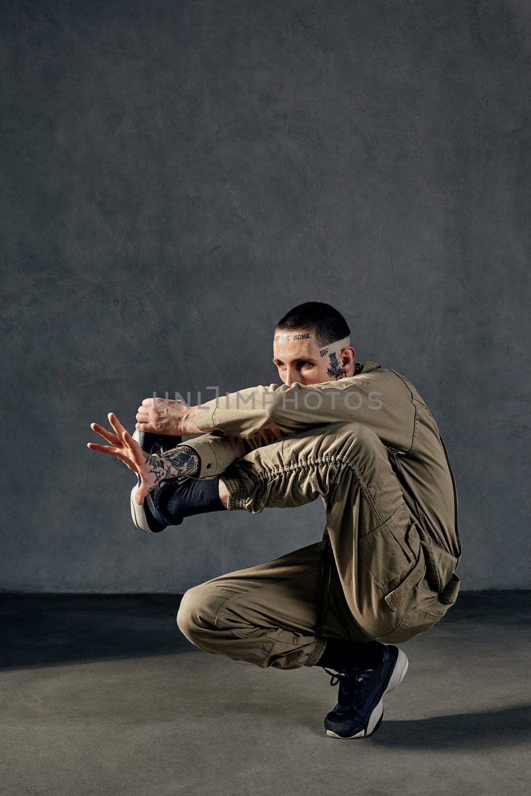 Young unusual fellow with tattooed body and face, earrings, beard. Dressed in khaki jumpsuit and black sneakers. He dancing against gray studio background. Dancehall, hip-hop. Full length, copy space