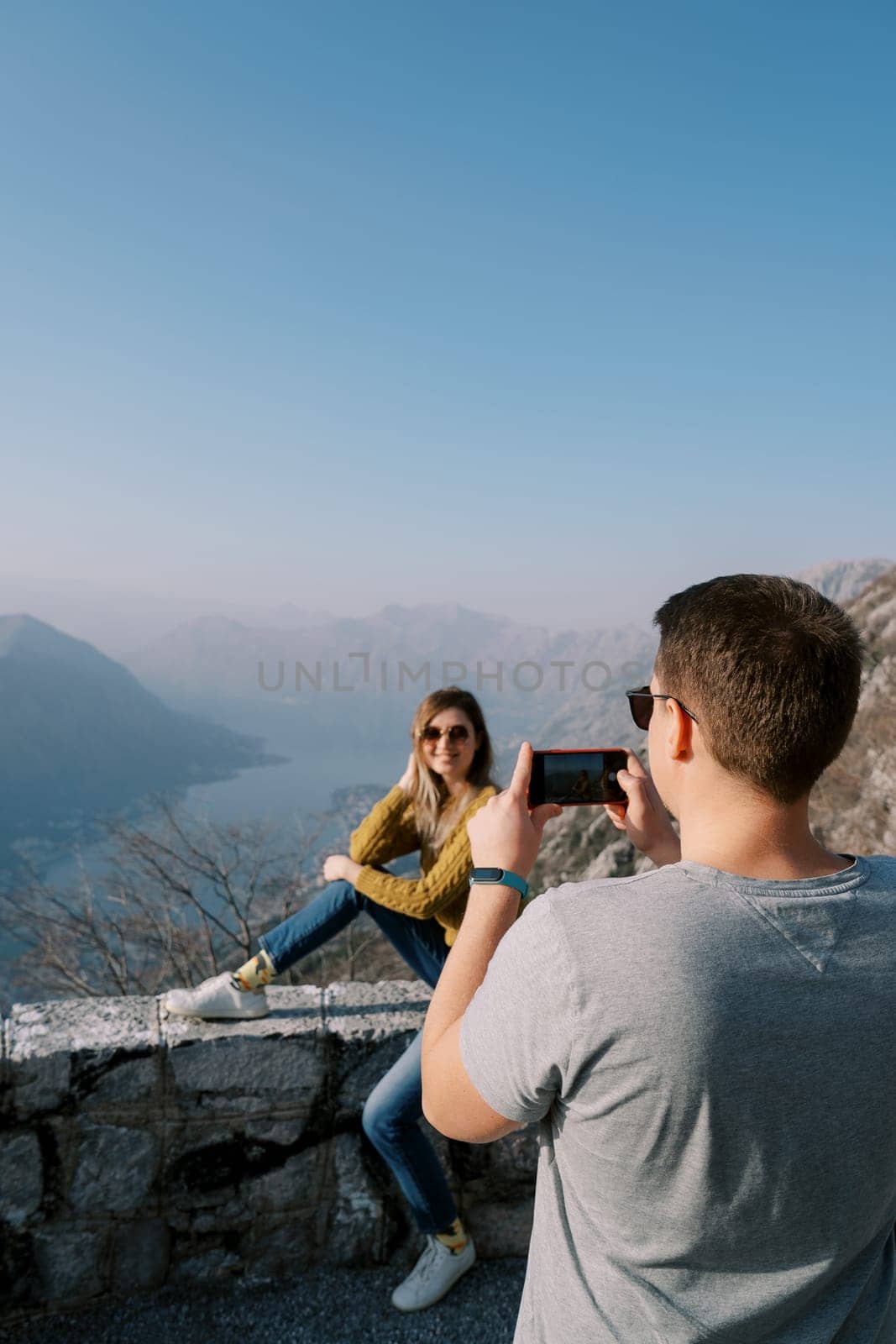 Man takes a picture with a smartphone of woman sitting on a fence over the Bay of Kotor. Montenegro. Back view by Nadtochiy