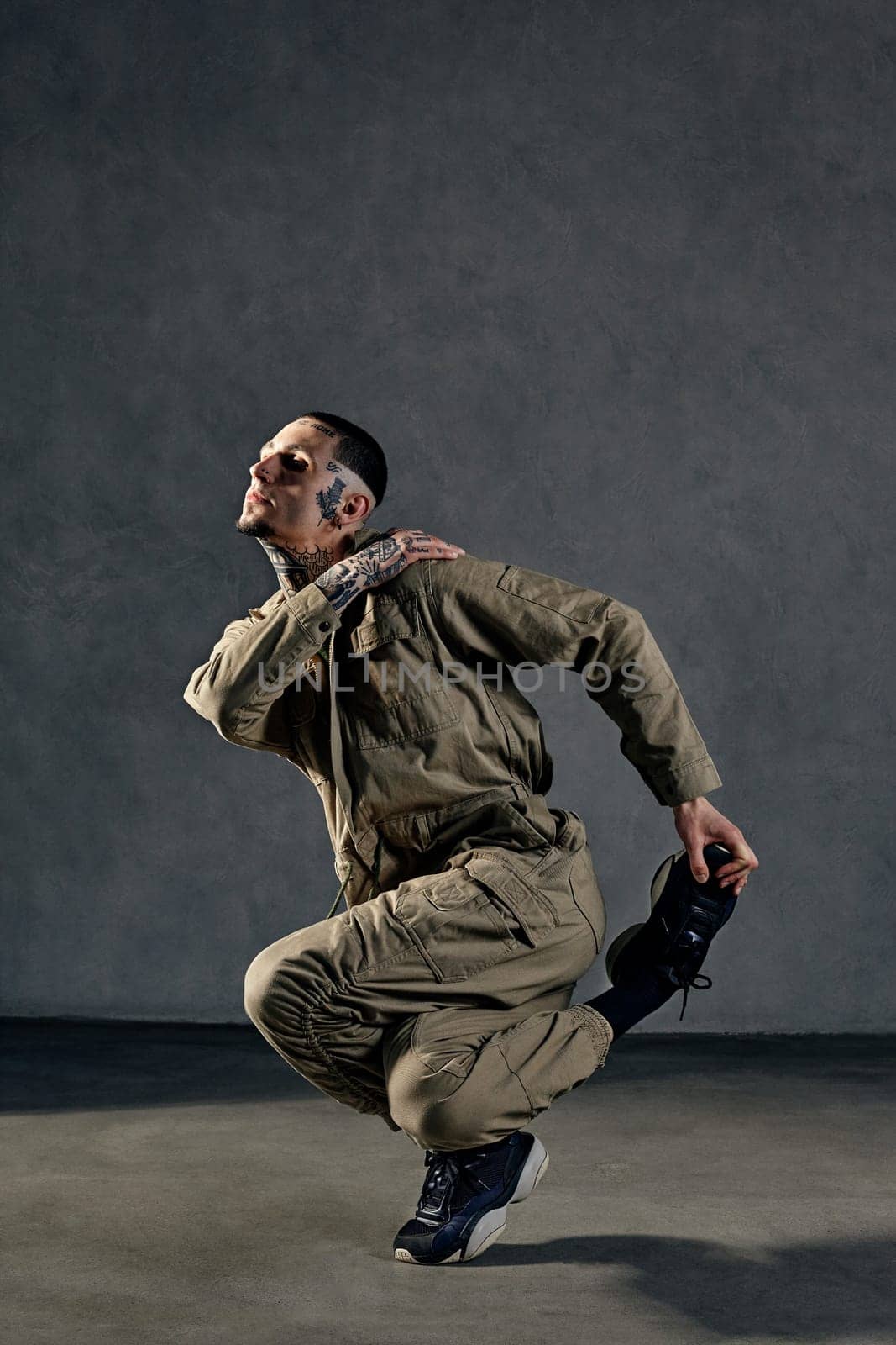 Extraordinary guy with tattooed body, earrings, beard. Dressed in khaki overalls and black sneakers. Dancing on gray background. Dancehall, hip-hop by nazarovsergey