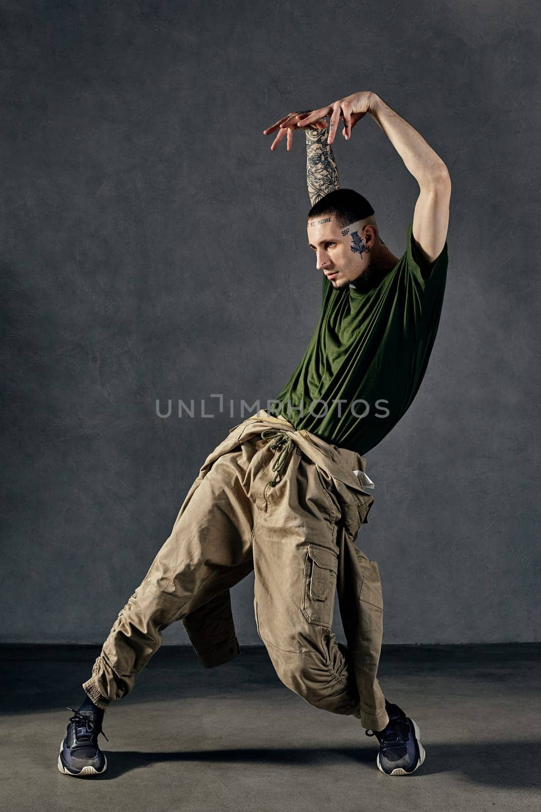 Young stylish fellow with tattooed body and face, earrings, beard. Dressed in khaki t-shirt and jumpsuit, black sneakers. Dancing against gray background. Dancehall, hip-hop. Full length, copy space