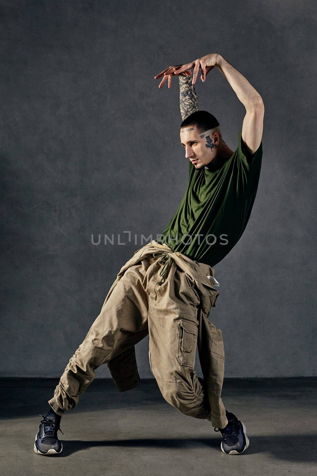 Fellow with tattooed body, earrings, beard. Dressed in khaki t-shirt and jumpsuit, black sneakers. Dancing on gray background. Dancehall, hip-hop by nazarovsergey