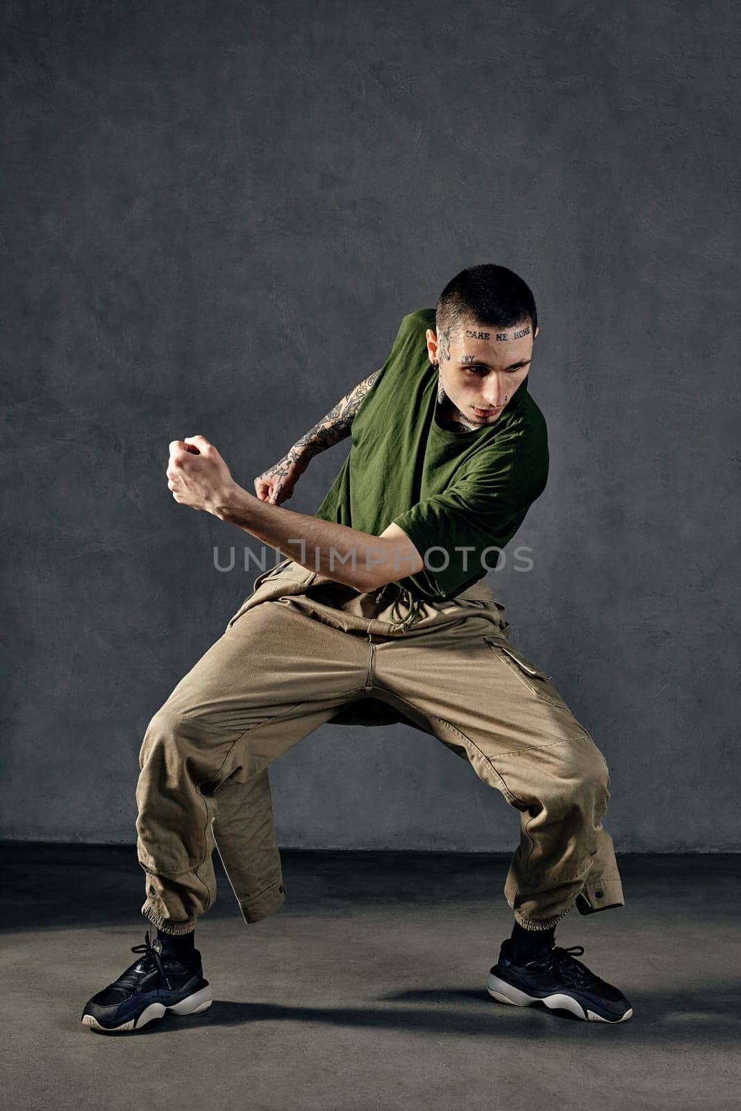 Young graceful male with tattooed body and face, earrings, beard. Dressed in khaki t-shirt and jumpsuit, black sneakers. Dancing against gray background. Dancehall, hip-hop. Full length, copy space