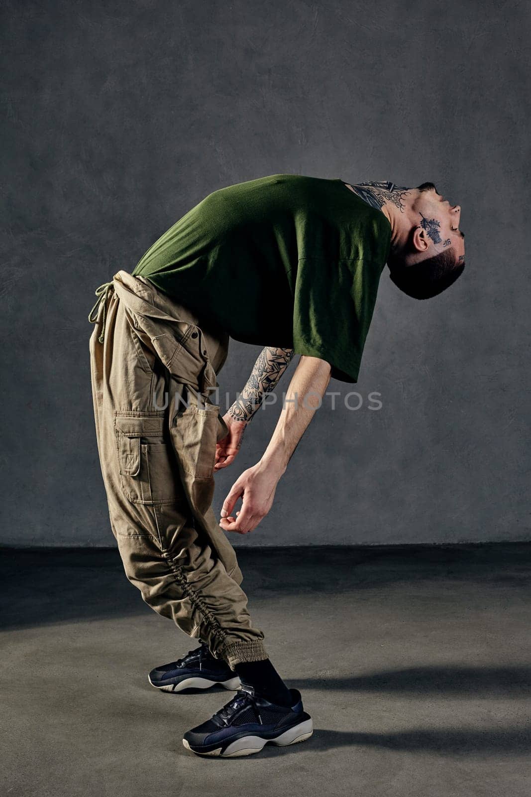 Graceful guy with tattooed body, earrings, beard. Dressed in khaki t-shirt, overalls, black sneakers. Dancing on gray background. Dancehall, hip-hop by nazarovsergey