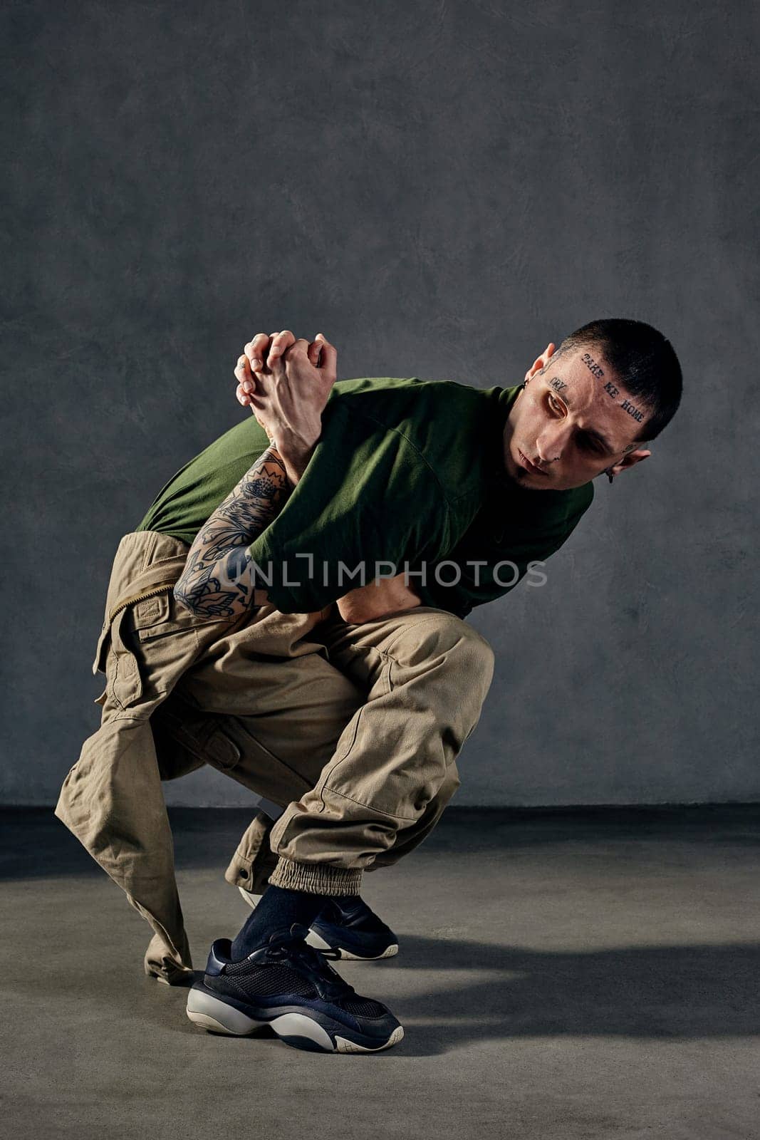 Graceful fellow with tattooed body, earrings, beard. Dressed in khaki t-shirt, jumpsuit, black sneakers. Dancing, gray background. Dancehall, hip-hop by nazarovsergey