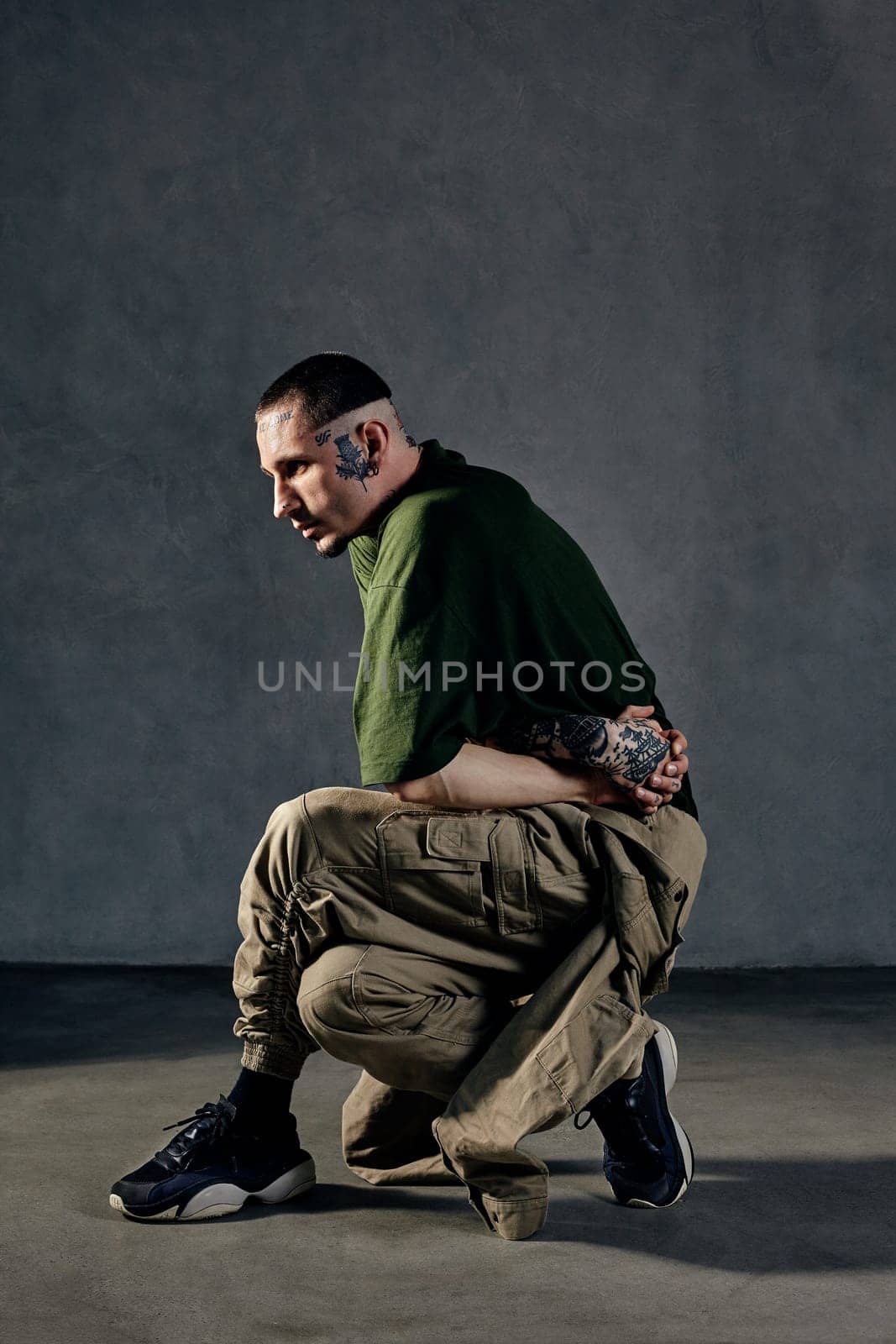 Young flexible man with tattooed body and face, earrings, beard. Dressed in green t-shirt and khaki overalls, black sneakers. Dancing on gray background. Dancehall, hip-hop. Full length, copy space