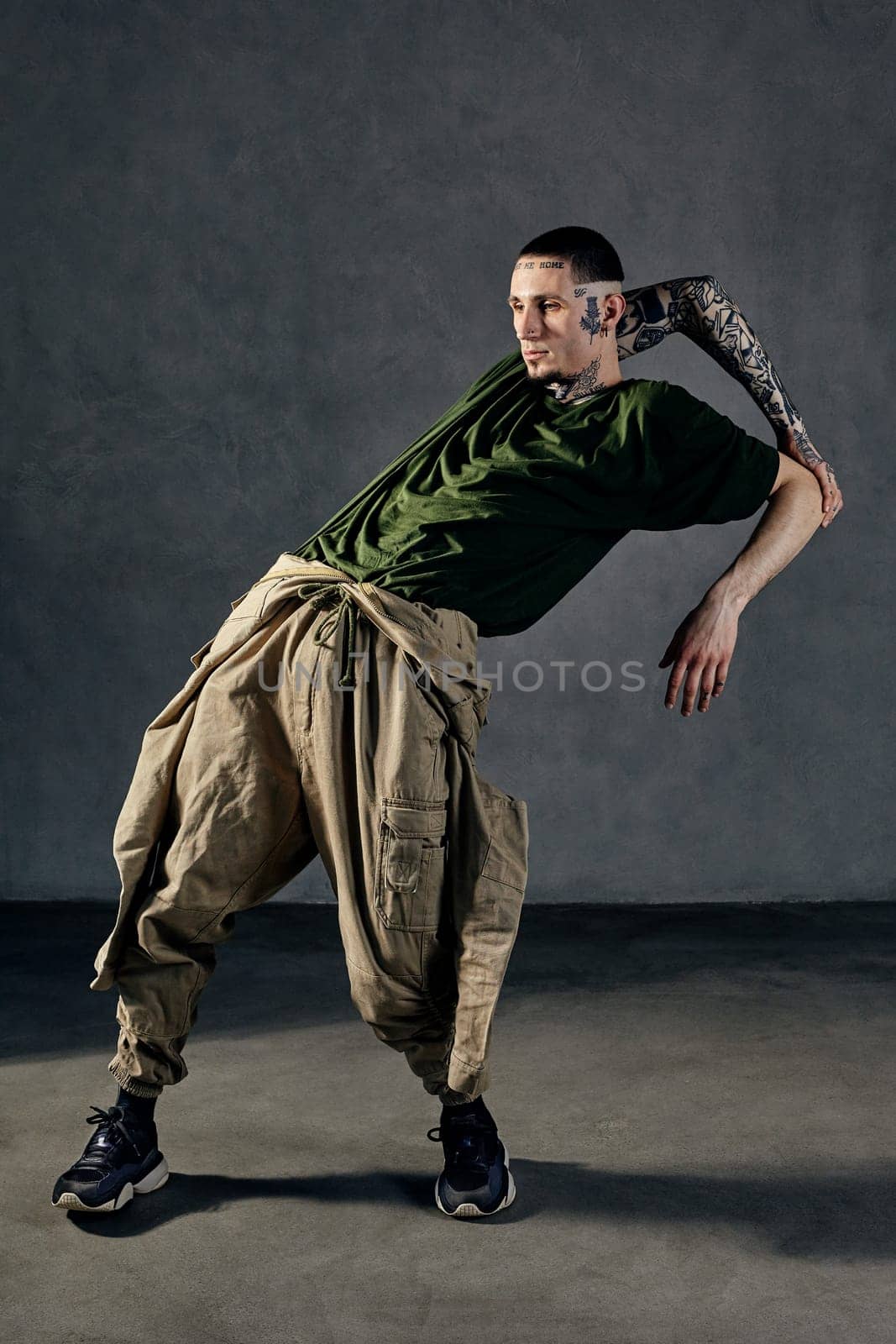 Young flexible guy with tattooed body and face, earrings, beard. Dressed in green t-shirt and khaki overalls, black sneakers. Dancing on gray background. Dancehall, hip-hop. Full length, copy space