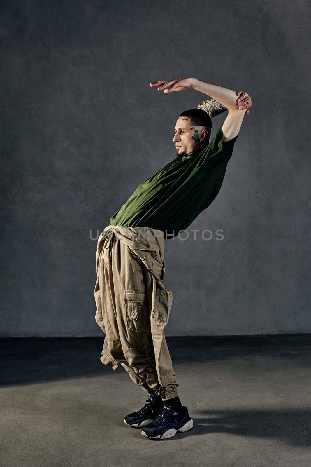 Young flexible fellow with tattooed body and face, earrings, beard. Dressed in khaki t-shirt and jumpsuit, black sneakers. Dancing against gray background. Dancehall, hip-hop. Full length, copy space