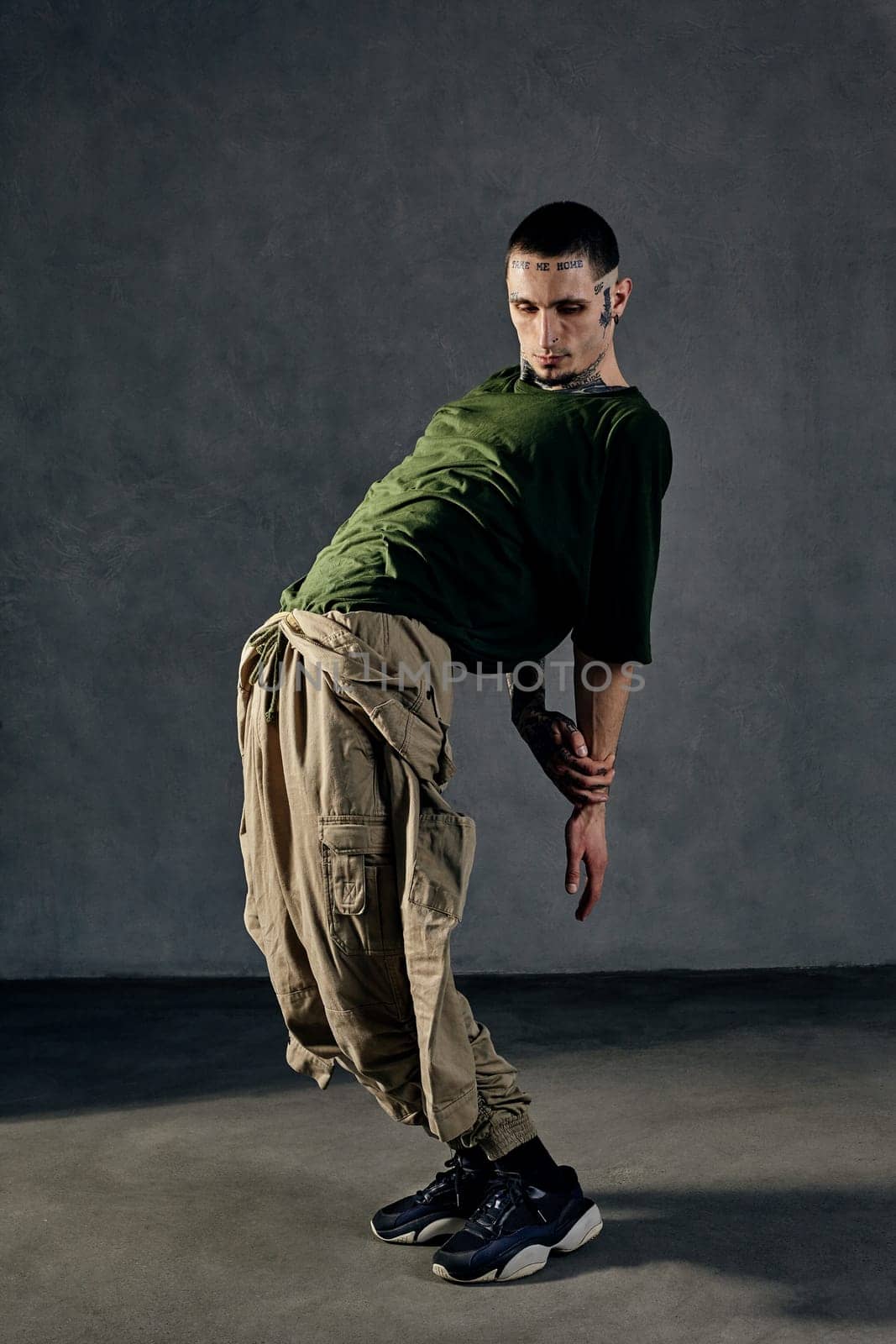 Young athletic male with tattooed body and face, earrings, beard. Dressed in khaki t-shirt and jumpsuit, black sneakers. Dancing against gray background. Dancehall, hip-hop. Full length, copy space