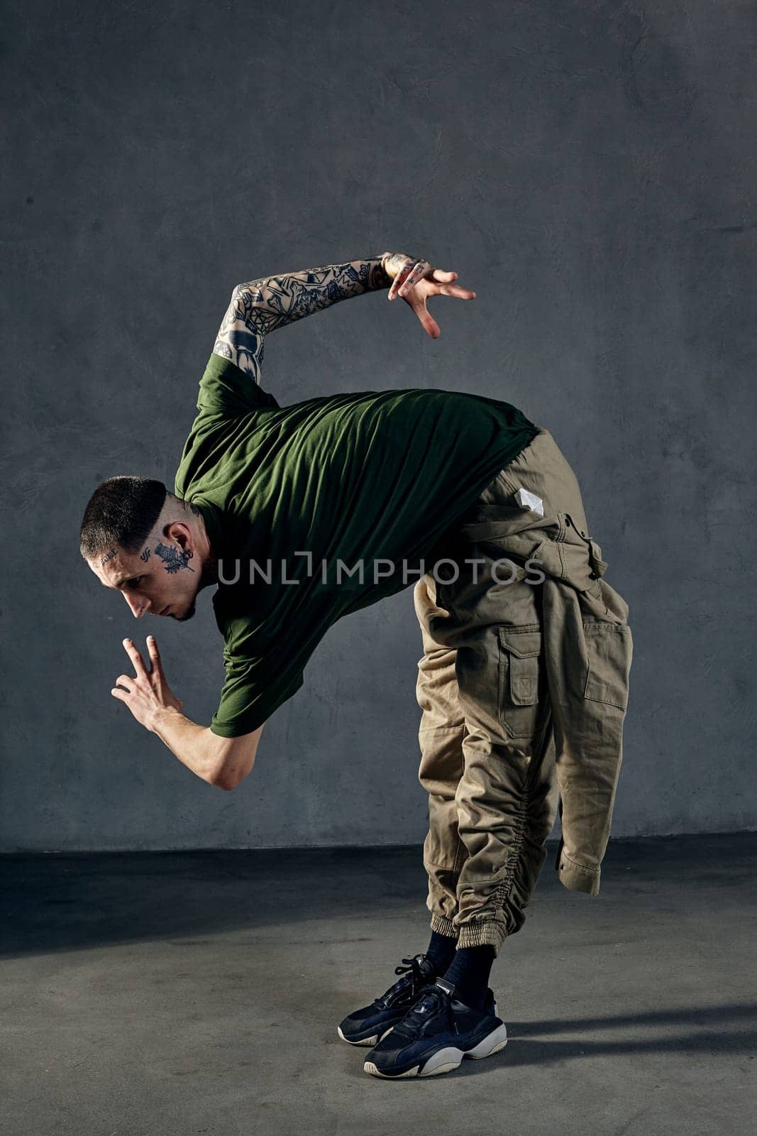 Young athletic guy with tattooed body and face, earrings, beard. Dressed in green t-shirt and khaki overalls, black sneakers. Dancing on gray background. Dancehall, hip-hop. Full length, copy space