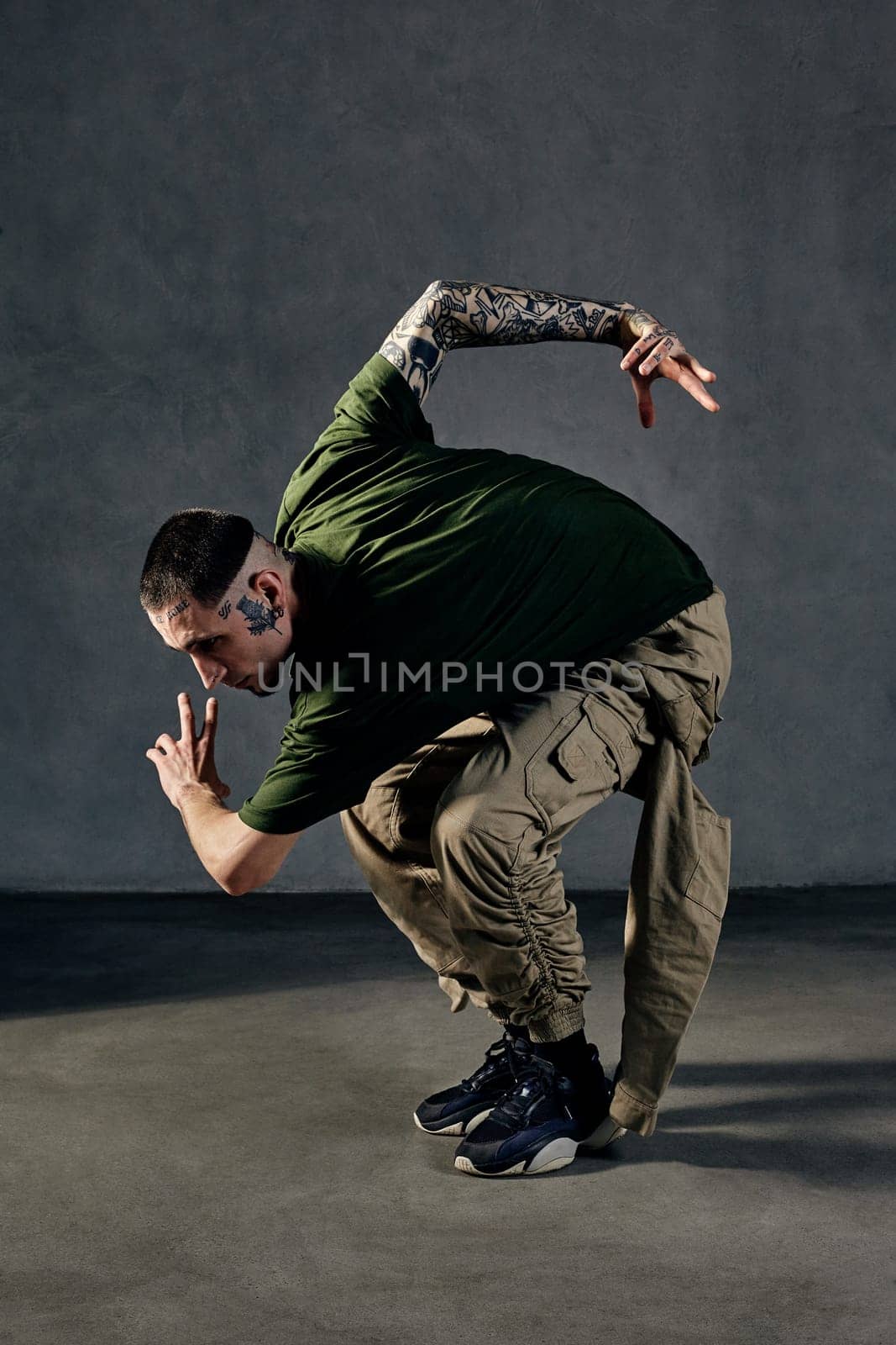 Athletic fellow with tattooed body, earrings, beard. Dressed in khaki t-shirt, jumpsuit, black sneakers. Dancing, gray background. Dancehall, hip-hop by nazarovsergey