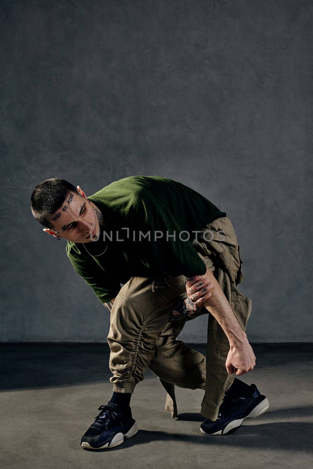 Young athletic performer with tattooed body and face, earrings, beard. Dressed in khaki t-shirt and overalls, black sneakers. Dancing on gray background. Dancehall, hip-hop. Full length, copy space