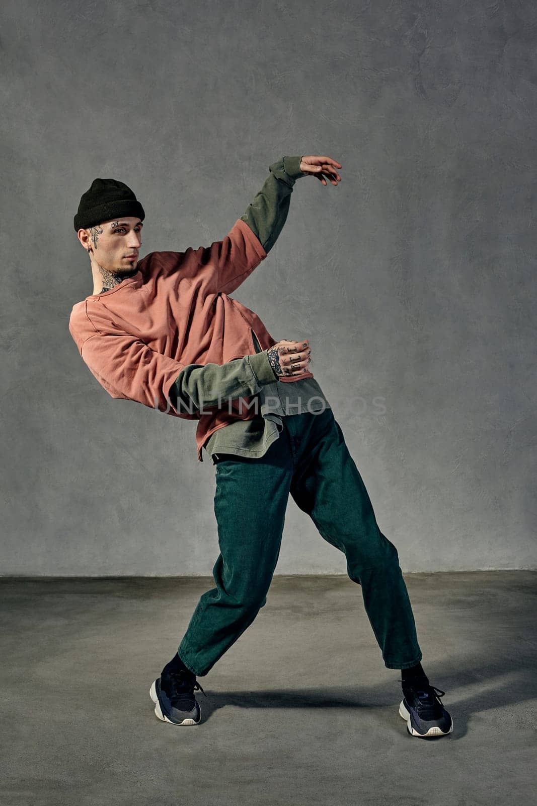 Young strong guy with tattooed body and face, earrings, beard. Dressed in hat, casual clothes and black sneakers. Dancing on gray background. Dancehall, hip-hop. Full length, copy space