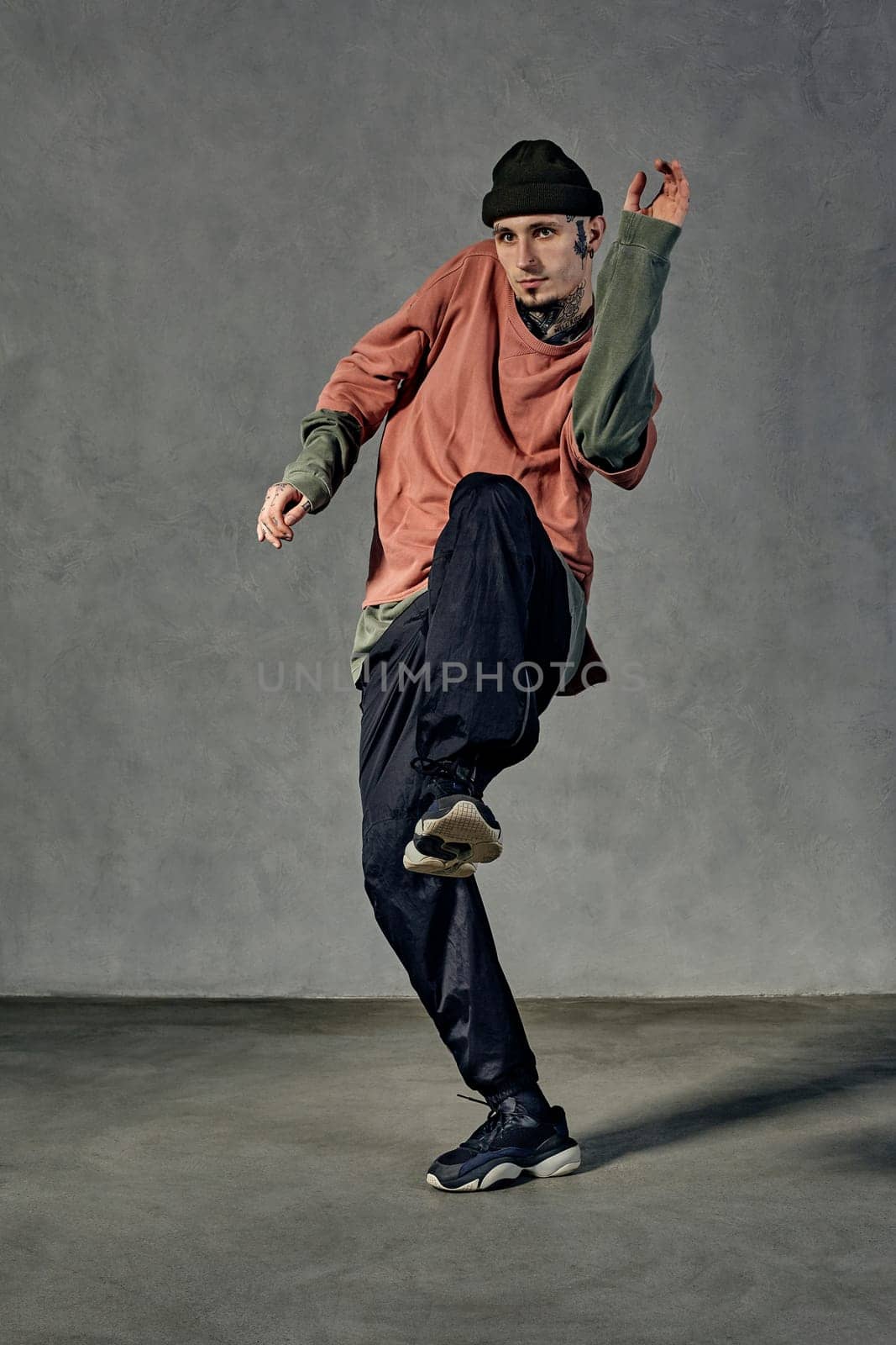 Strong fellow with tattooed body and face, earrings, beard. Dressed in hat, colorful jumper, black pants and sneakers. Dancing on gray background. Dancehall, hip-hop. Full length, copy space