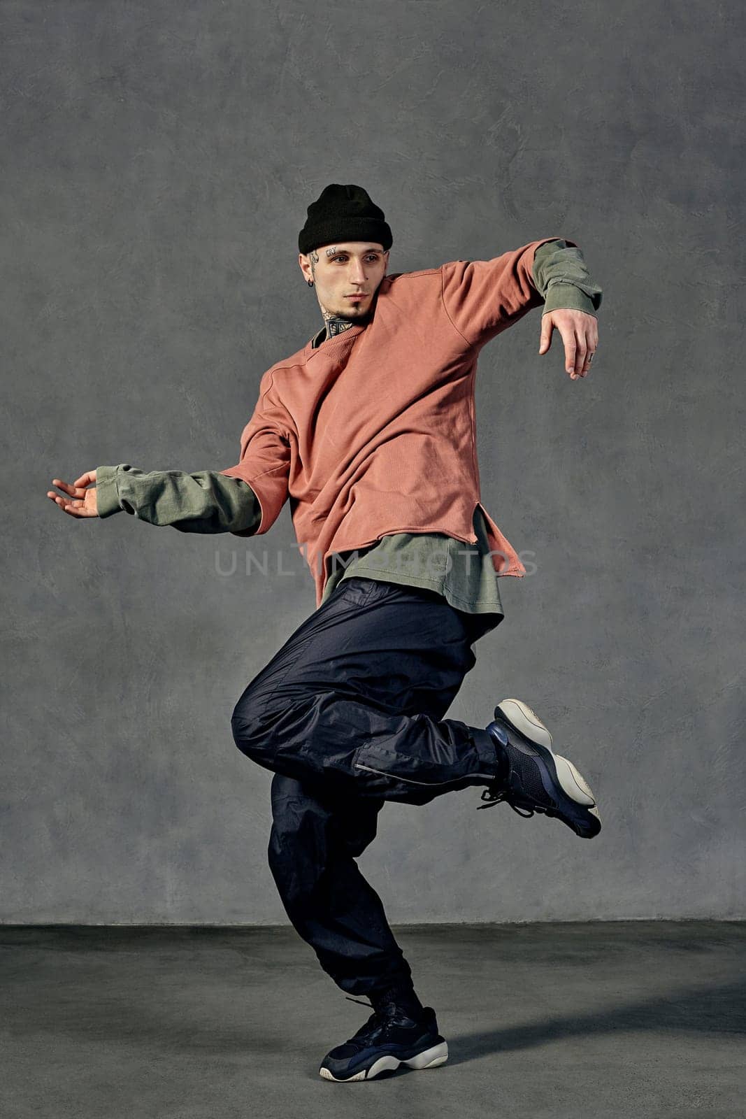 Young handsome man with tattooed body and face, earrings, beard. Dressed in hat, casual clothes and black sneakers. Dancing on gray background. Dancehall, hip-hop. Full length, copy space