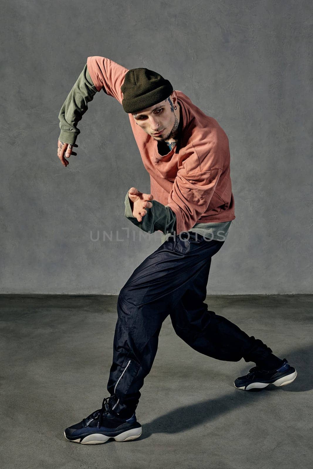 Young graceful guy with tattooed body and face, earrings, beard. Dressed in hat, casual clothes and black sneakers. Dancing on gray background. Dancehall, hip-hop. Full length, copy space