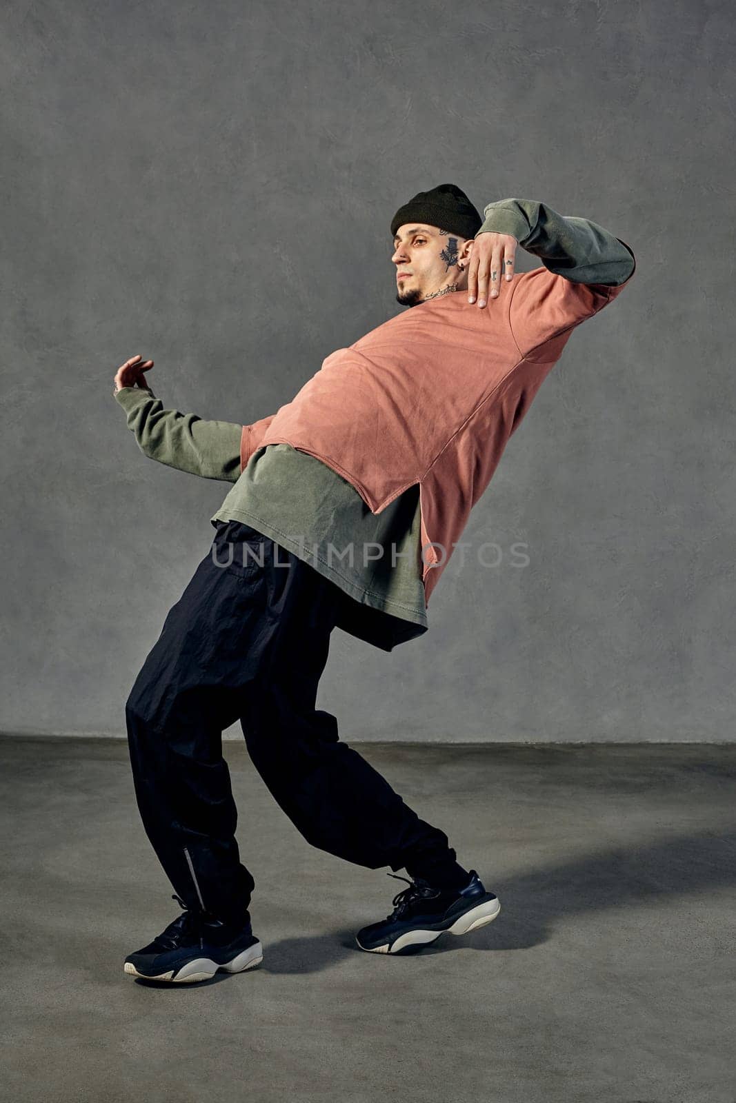 Graceful fellow with tattooed face, beard. Dressed in hat, colorful jumper, black pants and sneakers. Dancing, gray background. Dancehall, hip-hop by nazarovsergey