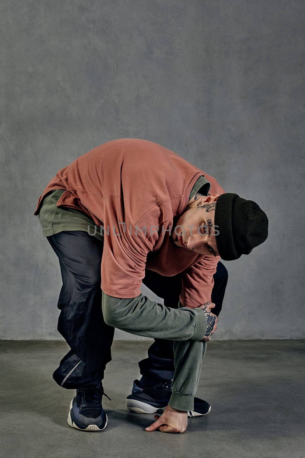 Modern male with tattooed body and face, earrings, beard. Dressed in colorful jumper, black pants and sneakers. Dancing on gray background. Dancehall, hip-hop. Full length, copy space