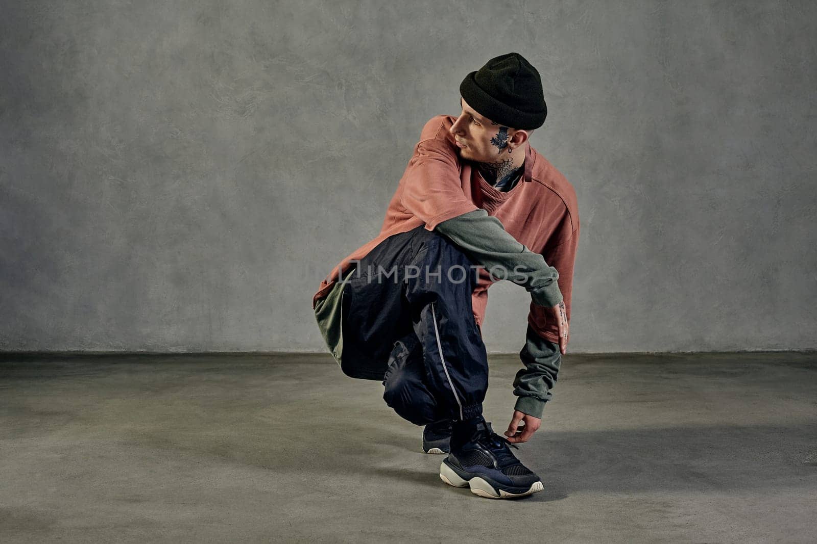 Young modern performer with tattooed body and face, earrings, beard. Dressed in hat, casual clothes and black sneakers. Dancing on gray background. Dancehall, hip-hop. Full length, copy space