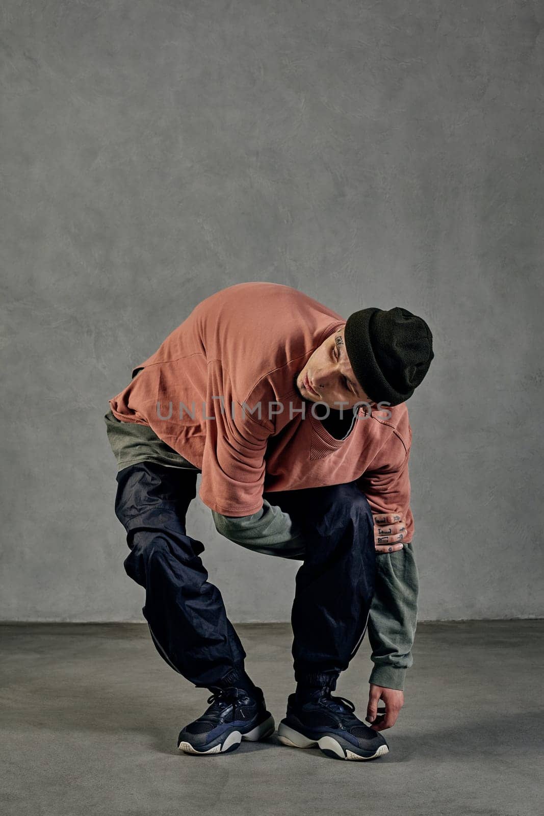 Young athletic man with tattooed body and face, earrings, beard. Dressed in hat, casual clothes and black sneakers. Dancing on gray background. Dancehall, hip-hop. Full length, copy space