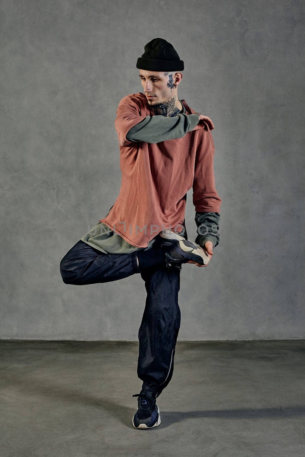 Athletic male with tattooed body and face, earrings, beard. Dressed in colorful jumper, black pants and sneakers. Dancing on gray background. Dancehall, hip-hop. Full length, copy space
