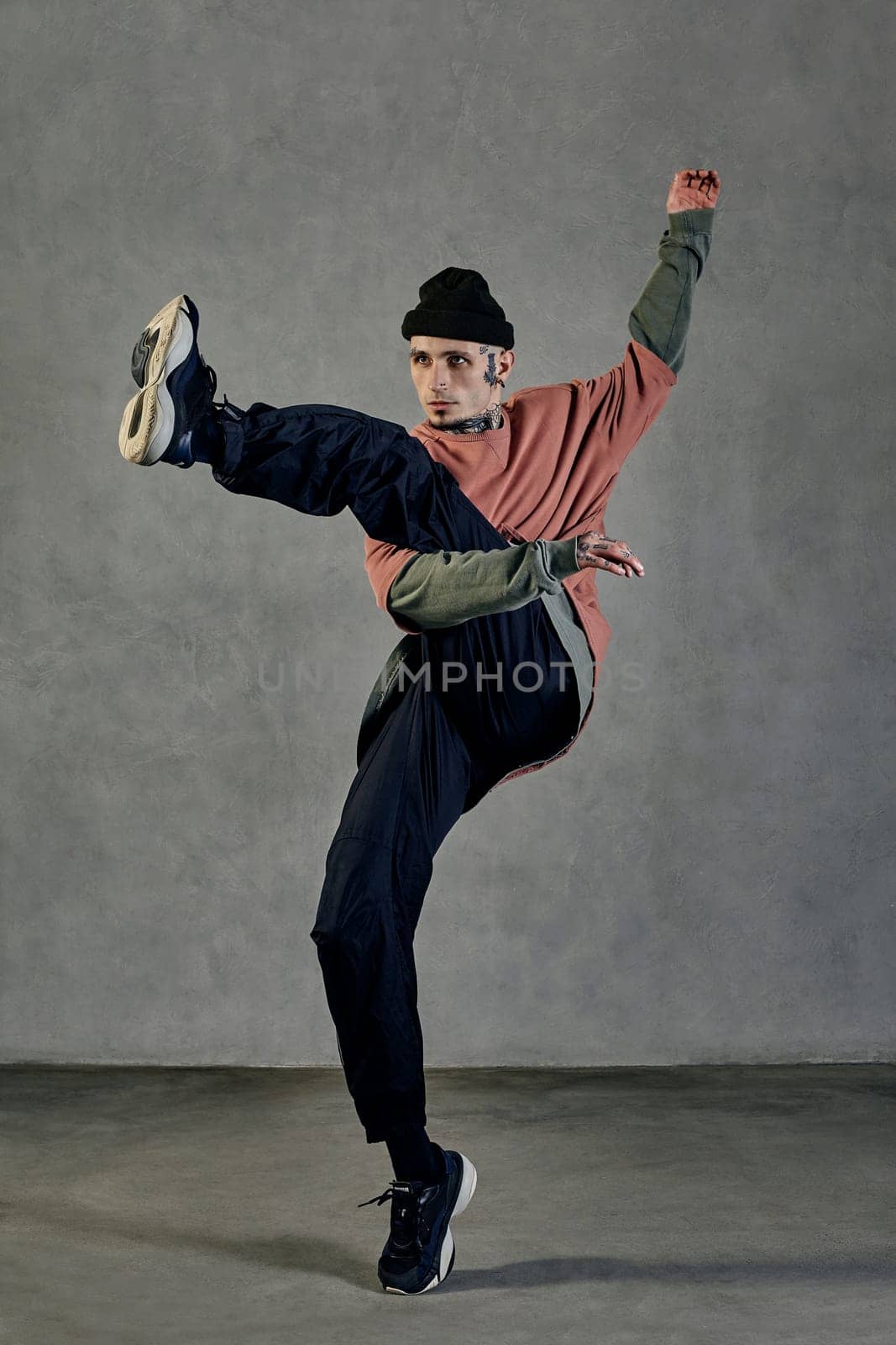 Athletic guy with tattooed body, earrings, beard. Dressed in hat, casual clothes and black sneakers. Dancing on gray background. Dancehall, hip-hop by nazarovsergey