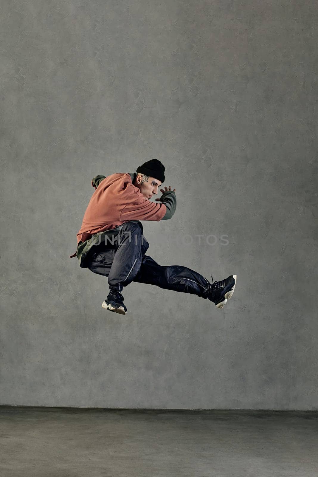 Young athletic performer with tattooed face, earrings, beard. Dressed in hat, casual clothes and black sneakers. Jumping while dancing on gray background. Dancehall, hip-hop. Full length, copy space