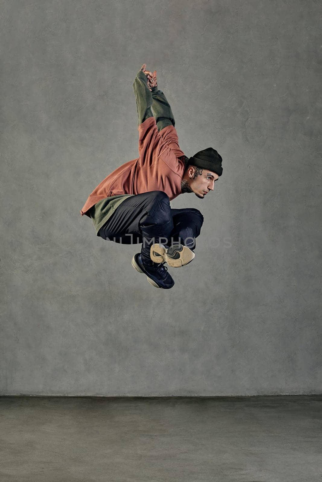 Young stately man with tattooed body and face, earrings, beard. Dressed in hat, casual clothes and black sneakers. Jumping while dancing on gray background. Dancehall, hip-hop. Full length, copy space