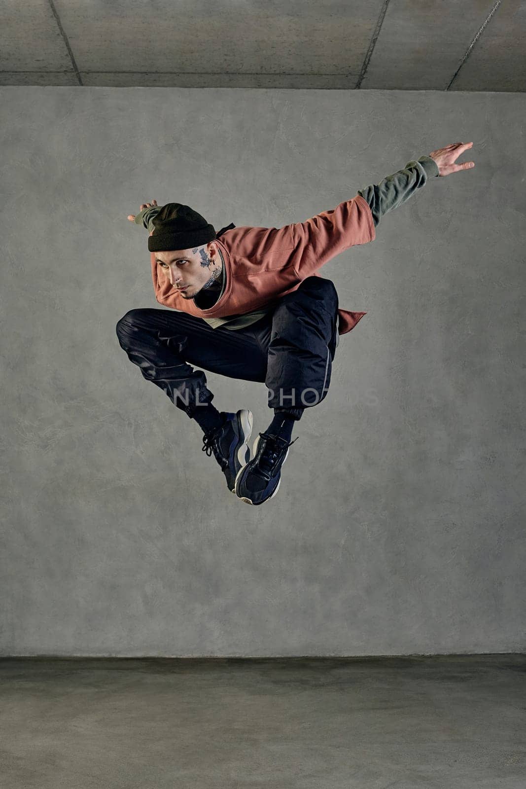 Stately guy with tattooed body, beard. Dressed in hat, casual clothes and black sneakers. Jumping while dancing on gray background. Dancehall, hip-hop by nazarovsergey