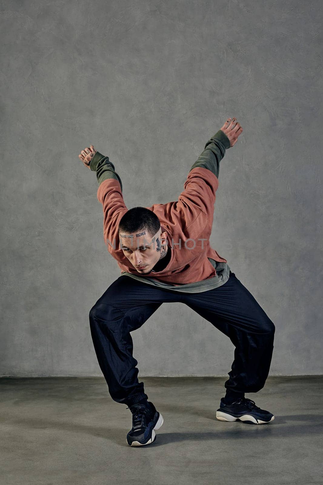 Young flexible man with tattooed body and face, earrings, beard. Dressed in casual clothes and black sneakers. Dancing on gray background. Dancehall, hip-hop. Full length, copy space