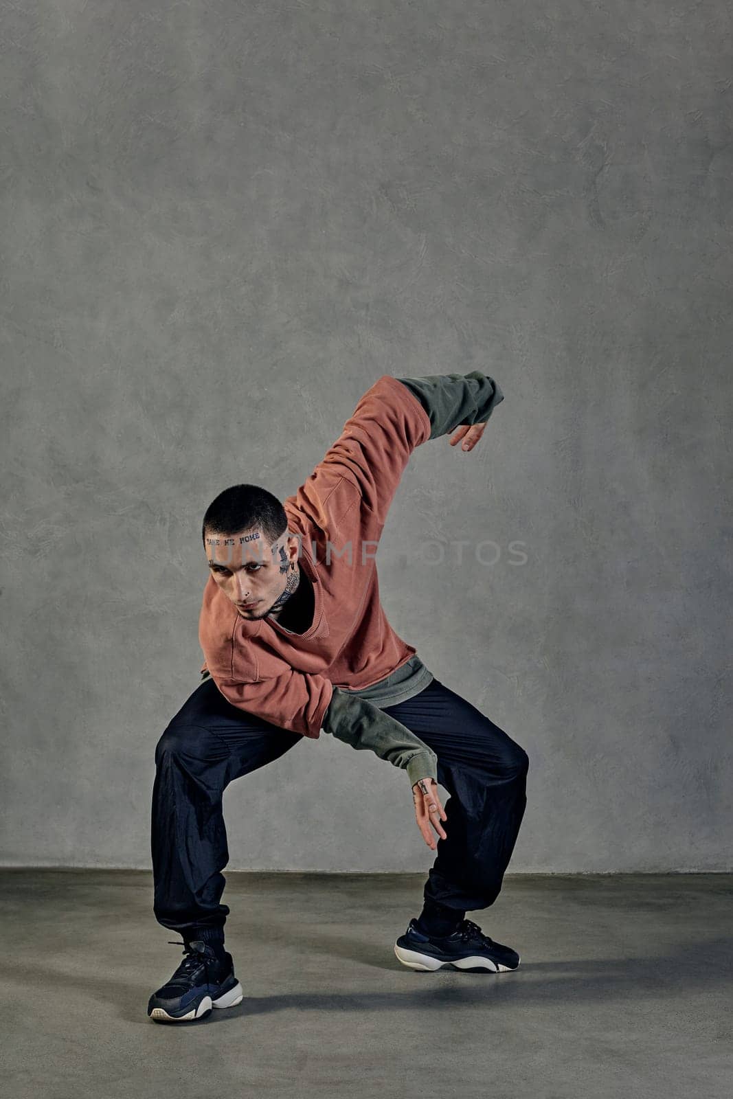 Flexible male with tattooed face, beard. Dressed in colorful jumper, black pants and sneakers. Dancing on gray background. Dancehall, hip-hop by nazarovsergey