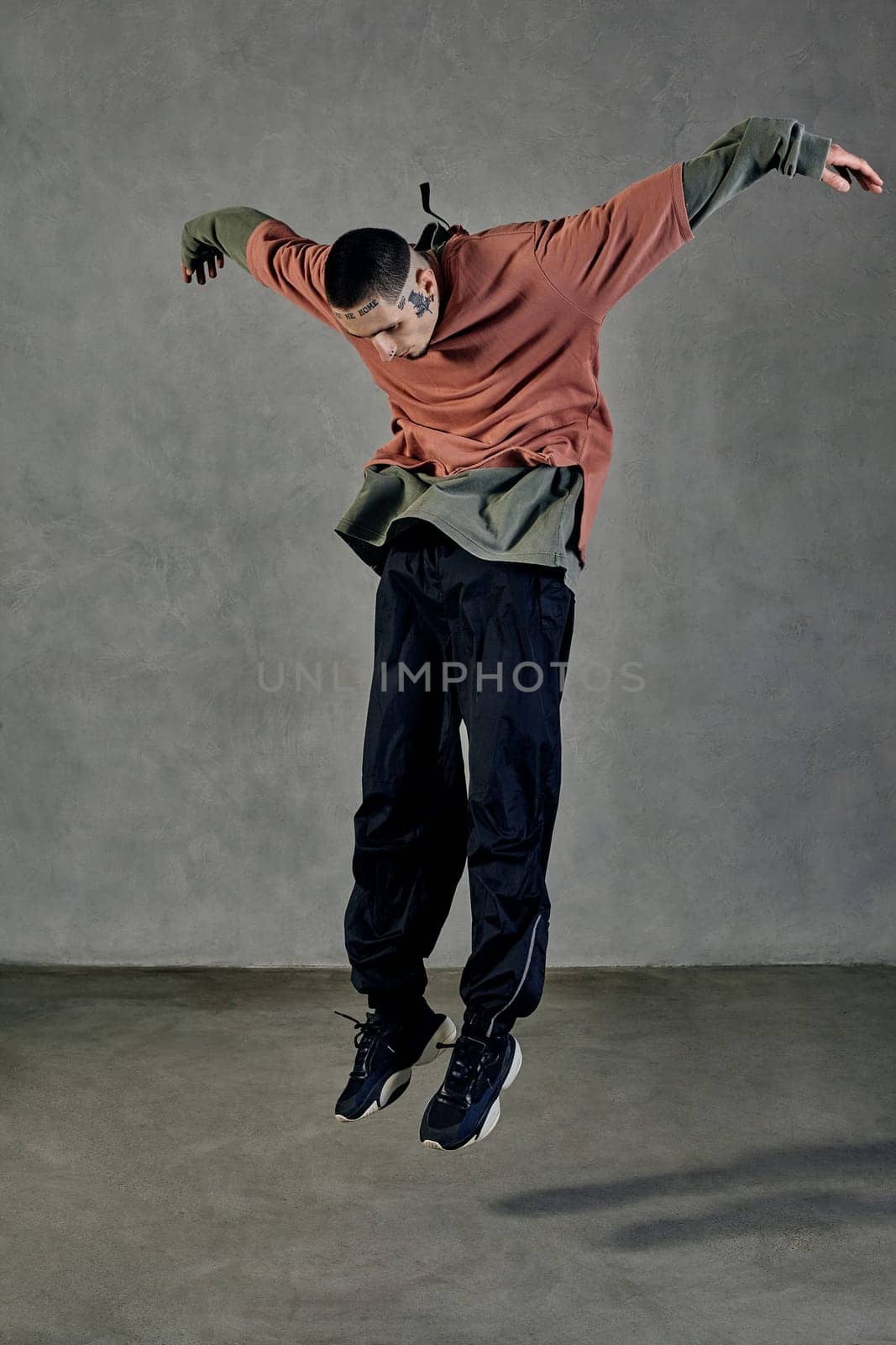 Young flexible guy with tattooed body and face, earrings, beard. Dressed in casual clothes and black sneakers. Jumping while dancing on gray background. Dancehall, hip-hop. Full length, copy space