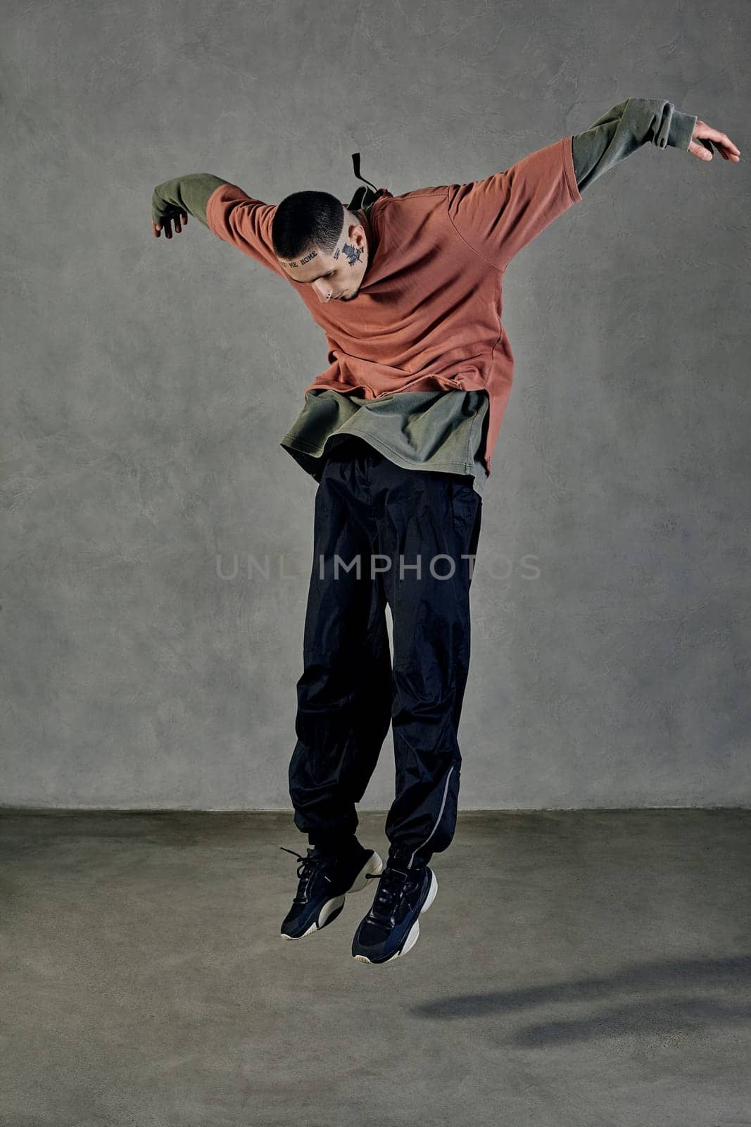 Flexible guy with tattooed body, beard. Dressed in casual clothes and black sneakers. Jumping while dancing on gray background. Dancehall, hip-hop by nazarovsergey