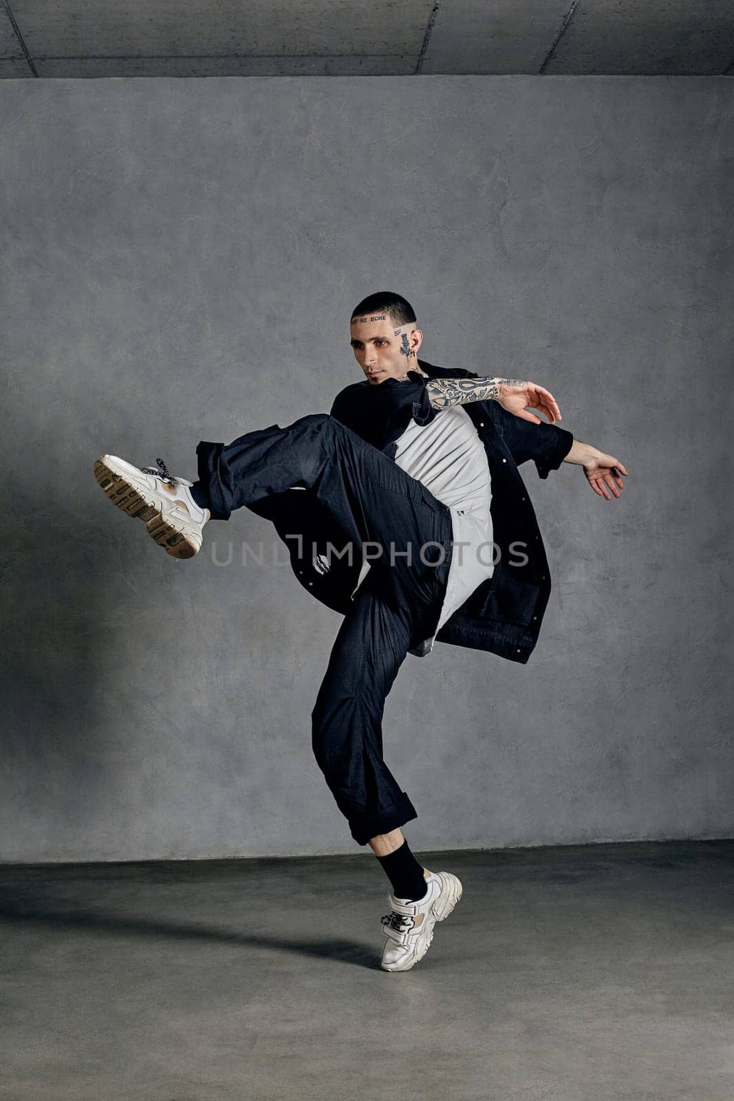 Graceful performer, tattooed body, beard. Dressed in white t-shirt and sneakers, black shirt, pants. Dancing on gray background. Dancehall, hip-hop by nazarovsergey