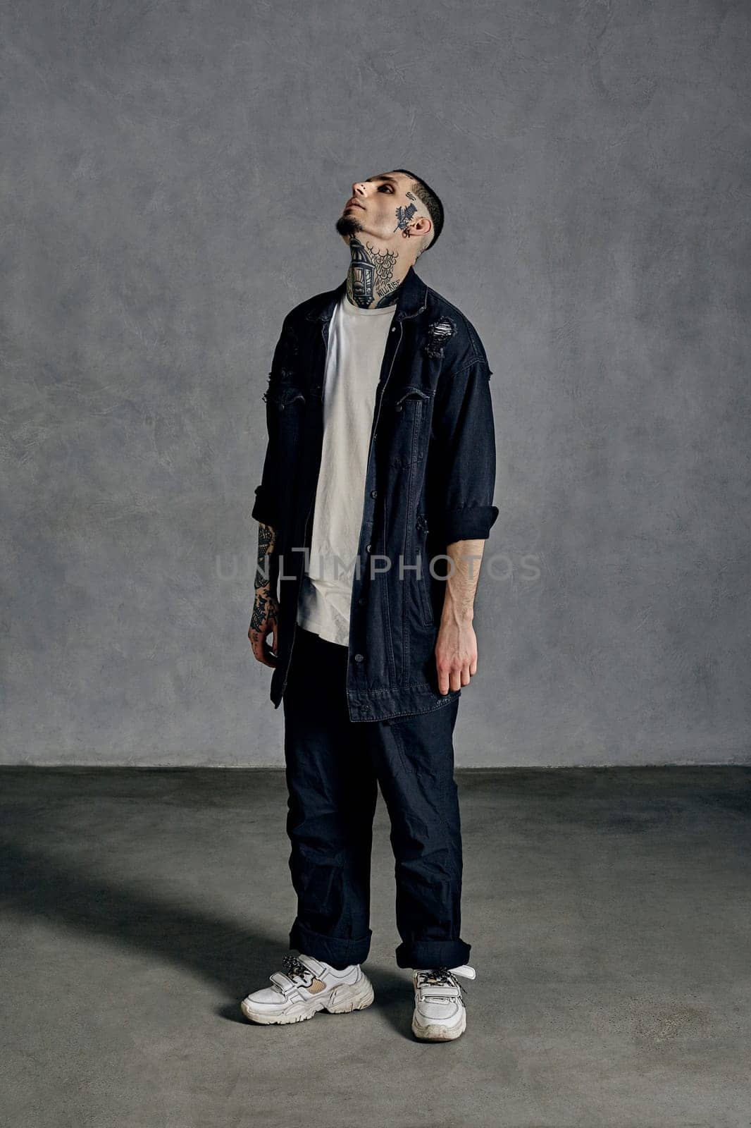 Athletic male with tattooed body and face, beard. Dressed in white t-shirt and sneakers, black denim shirt, pants. Looking up, standing against gray background. Dancehall, hip-hop. Full length