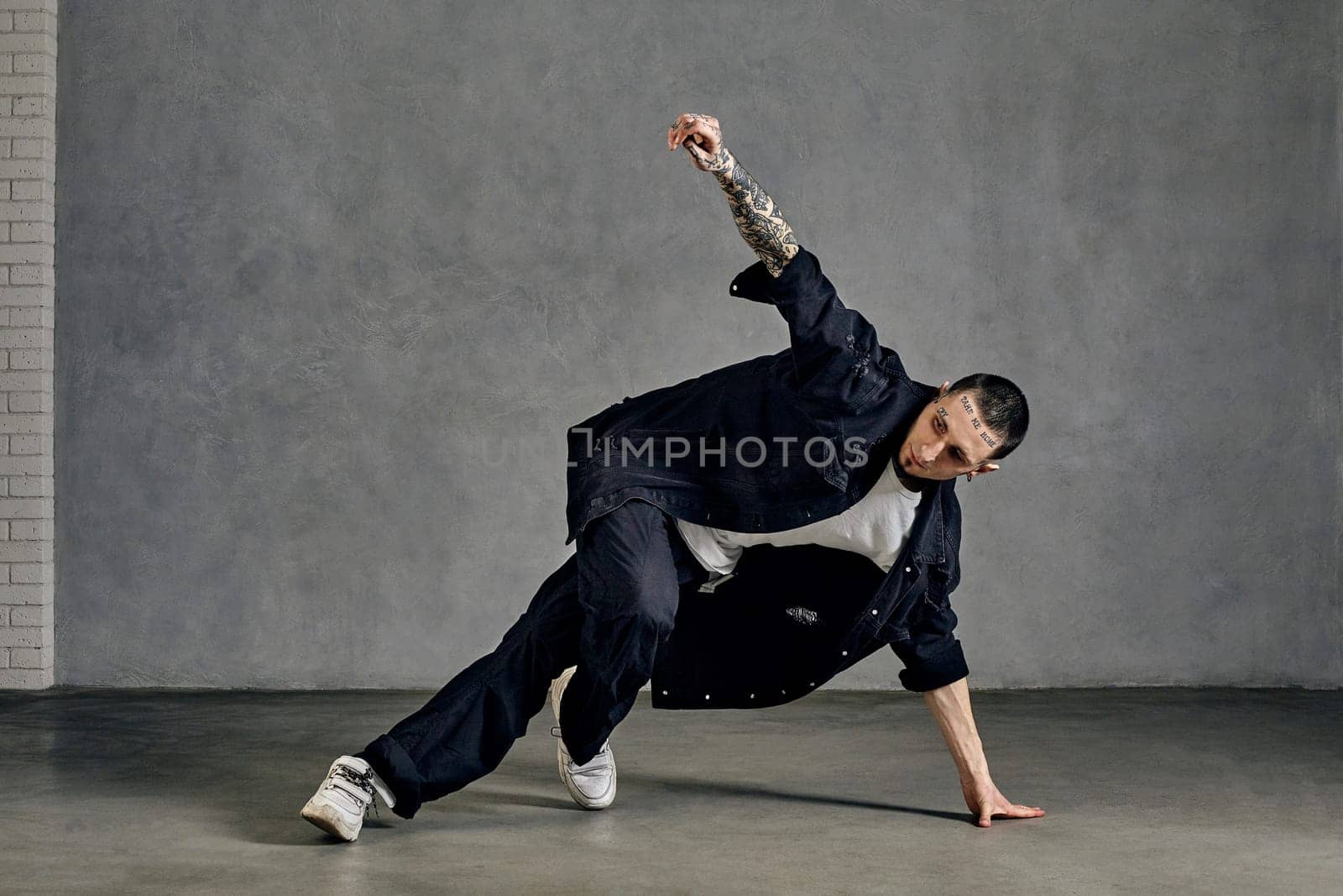 Athletic performer with tattooed body, earrings, beard. Dressed in white t-shirt and sneakers, black denim shirt and pants. Dancing on gray background. Dancehall, hip-hop. Full length, copy space