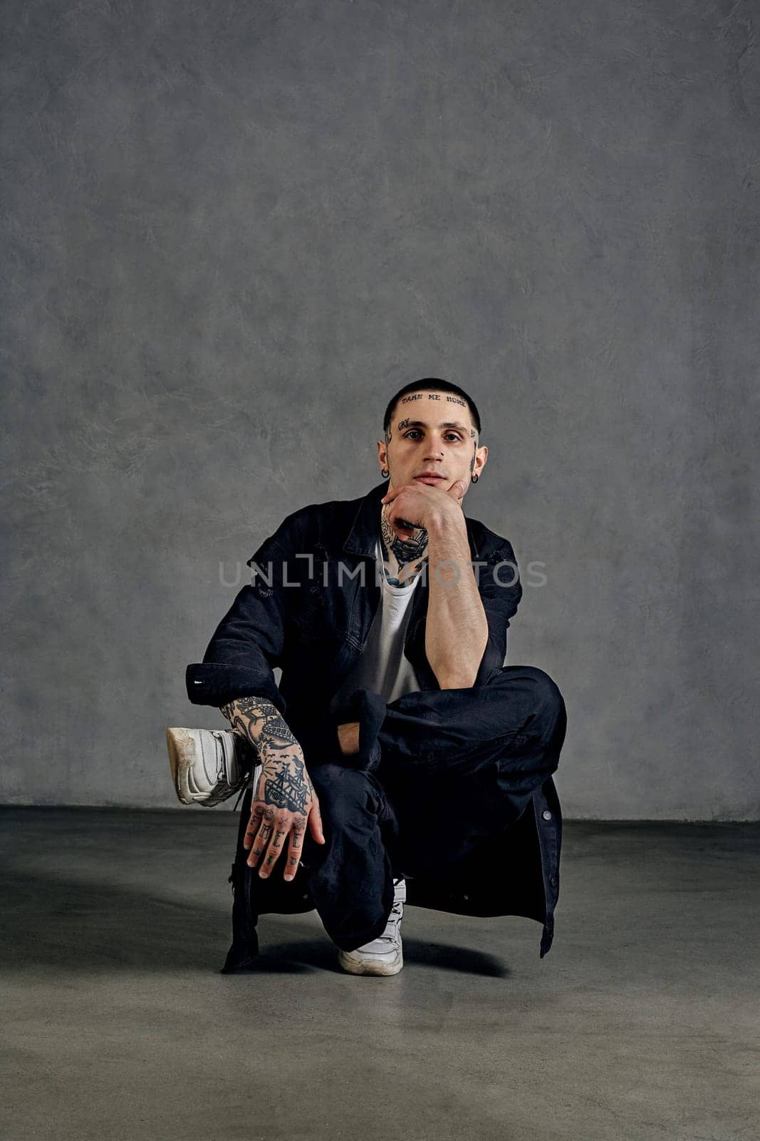 Stylish man with tattooed body, earrings, beard. Dressed in white t-shirt and sneakers, black denim shirt and pants. Performing tricks on gray background. Dancehall, hip-hop. Close up, copy space