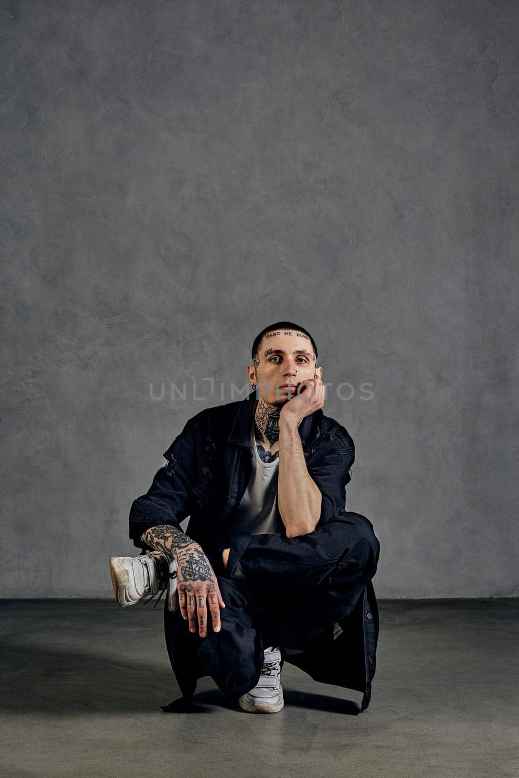 Stylish male with tattooed body and face, earrings, beard. Dressed in white t-shirt and sneakers, black denim shirt, pants. Squatting against gray background. Dancehall, hip-hop. Close up