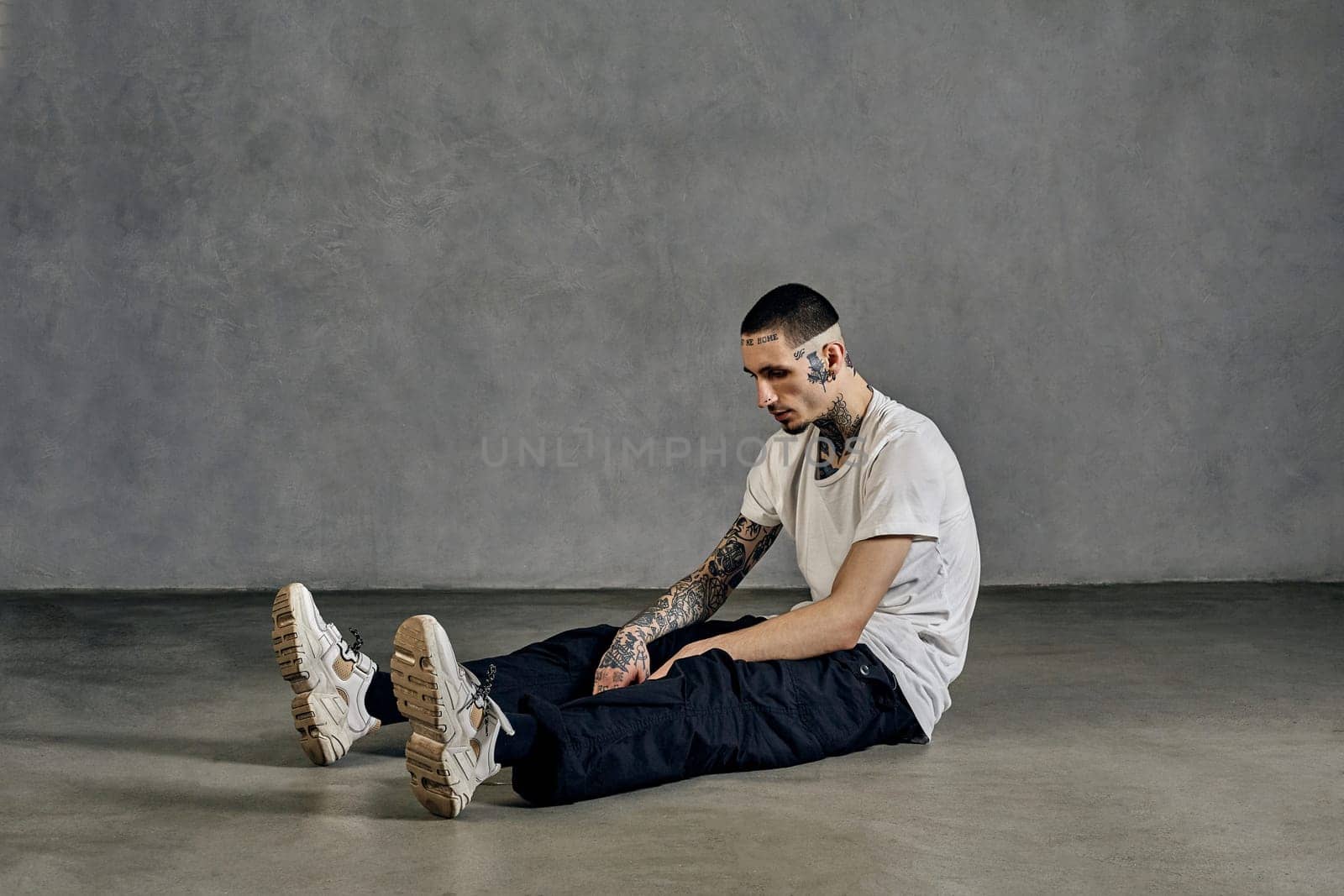 Athletic tattooed guy with beard. Dressed in white t-shirt, sneakers, black sports trousers. Sitting on floor, gray background. Dancehall, hip-hop by nazarovsergey