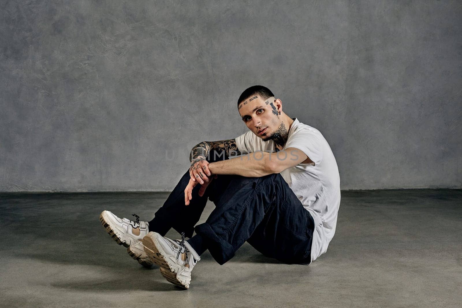 Athletic fellow with tattooed body and face, beard. Dressed in white t-shirt and sneakers, black sports trousers. Sitting sideways on floor against gray background. Dancehall, hip-hop. Close up