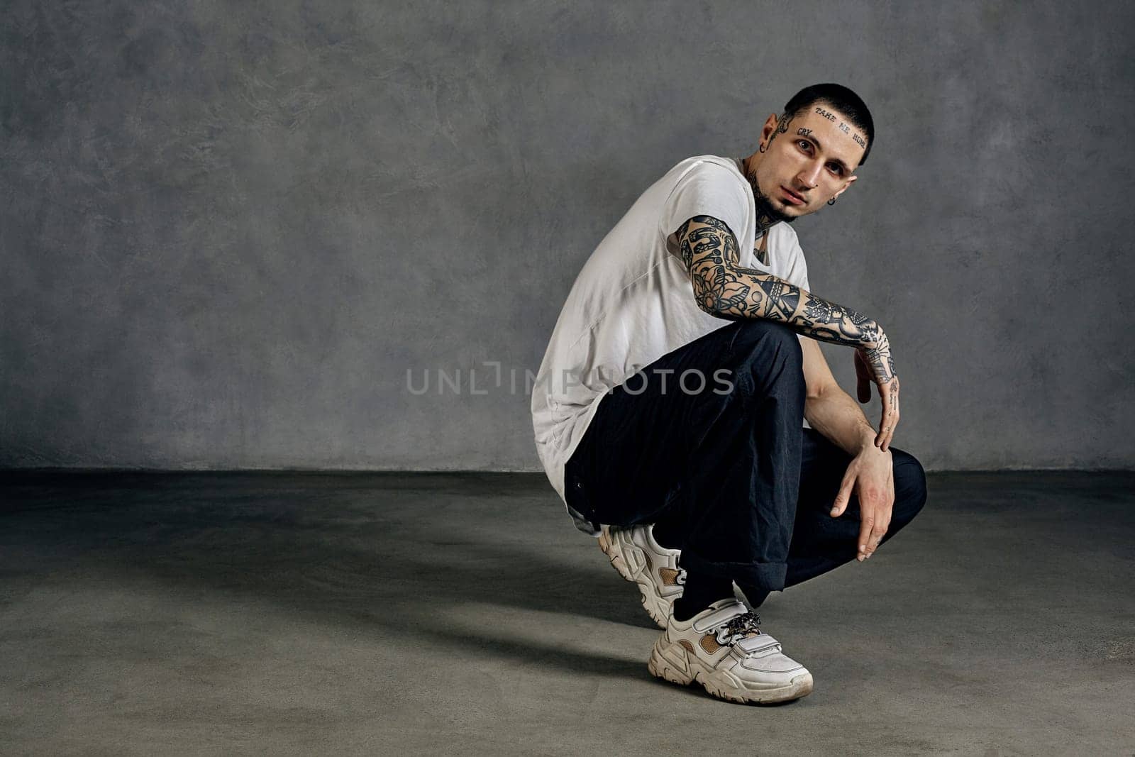 Flexible guy with tattooed body and face, earrings, beard. Dressed in white t-shirt and sneakers, black sports trousers. Squatting sideways against gray background. Dancehall, hip-hop. Close up