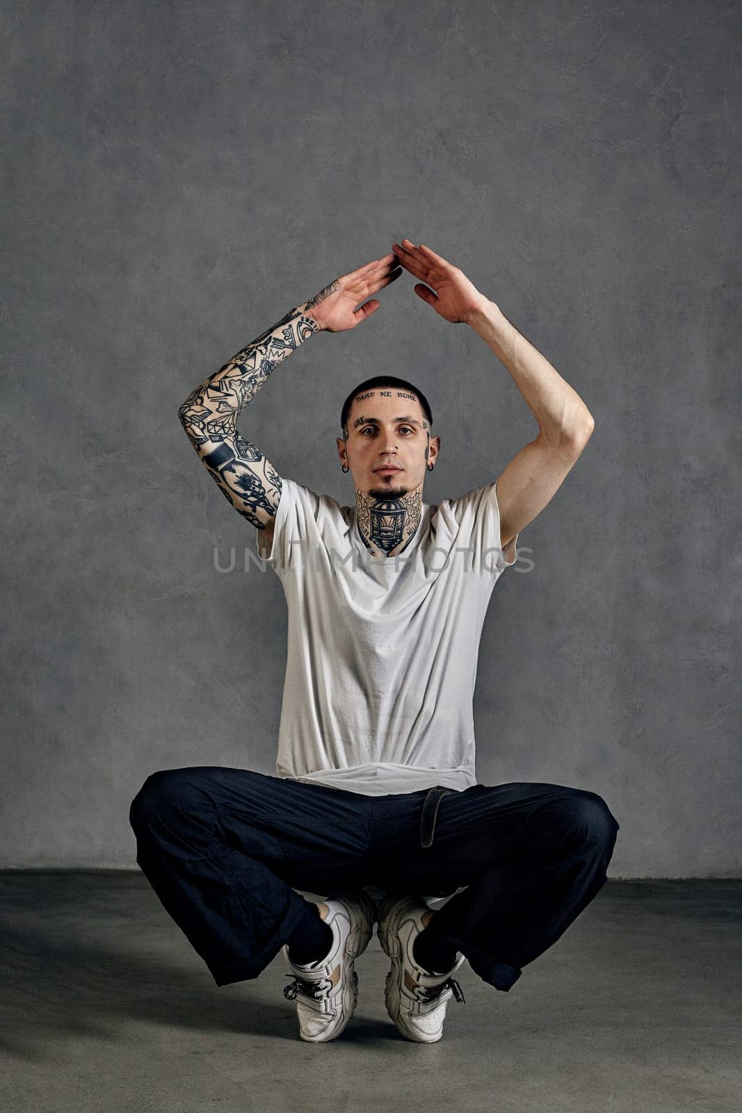 Flexible fellow with tattooed body and face, beard. Dressed in white t-shirt and sneakers, black sports trousers. Performing tricks against gray background. Dancehall, hip-hop. Close up, copy space