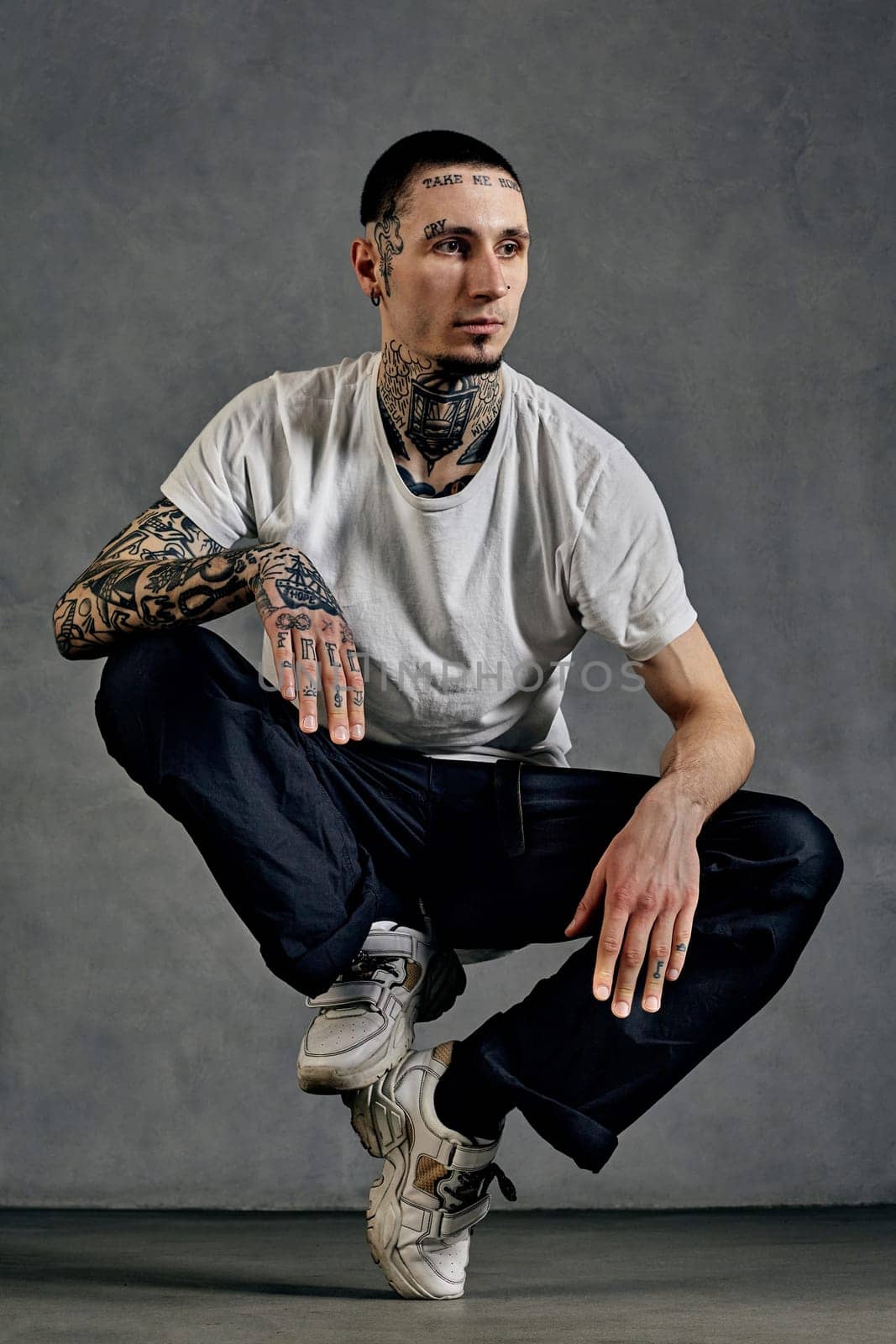 Strong male with tattooed body and face, beard. Dressed in white t-shirt and sneakers, black sports trousers. Performing tricks against gray background. Dancehall, hip-hop. Close up, copy space