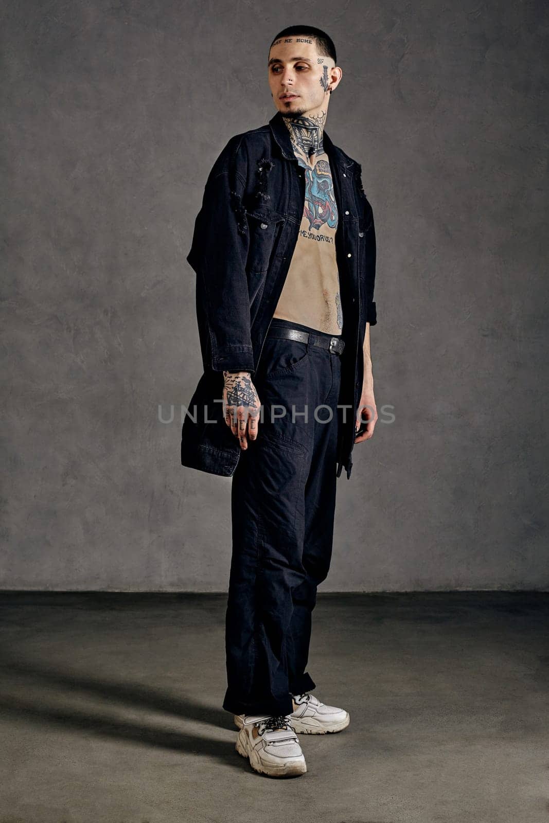 Strong guy with tattooed body and face, naked torso, beard. Dressed in black denim shirt, pants and white sneakers. Looking aside, posing on gray background. Dancehall, hip-hop. Full length