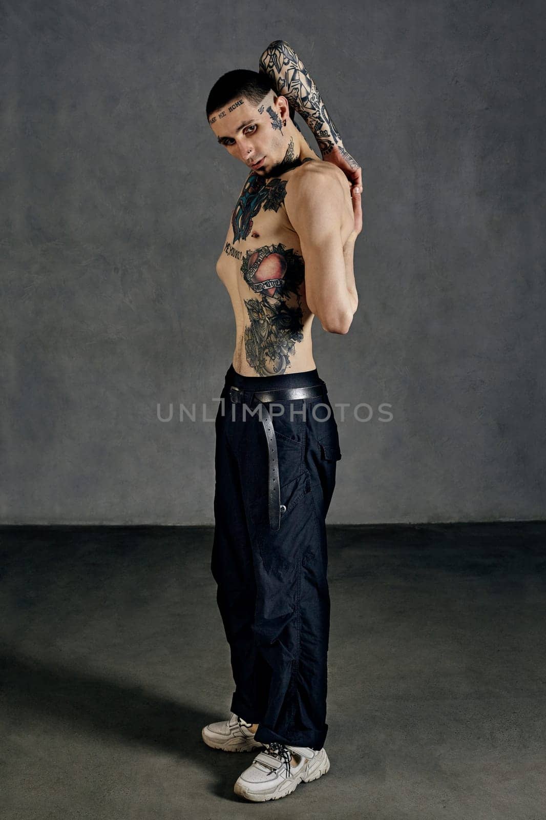 Strong fellow with tattooed body and face, naked torso, beard. Dressed in black pants and white sneakers. Stretching while posing sideways against gray background. Dancehall, hip-hop. Full length