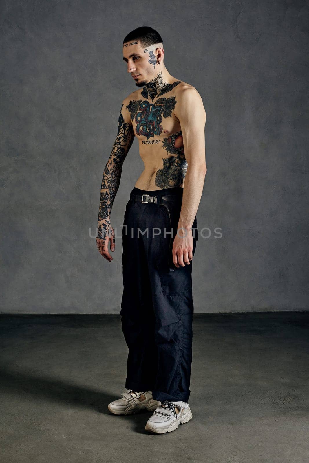 Strong performer with tattooed body and face, naked torso, beard. Dressed in black pants and white sneakers. He is dancing against gray studio background. Dancehall, hip-hop. Full length, copy space