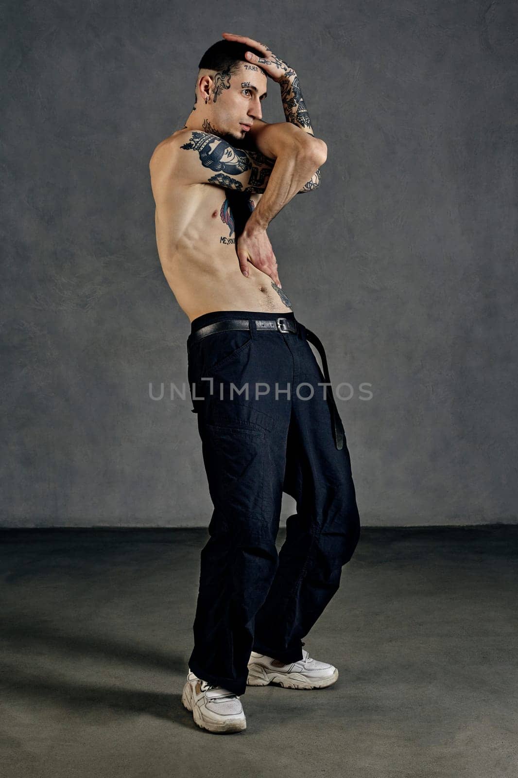 Athletic tattooed man with naked torso, beard. Dressed in black pants and white sneakers. He is dancing against gray studio background. Dancehall, hip-hop. Full length, copy space