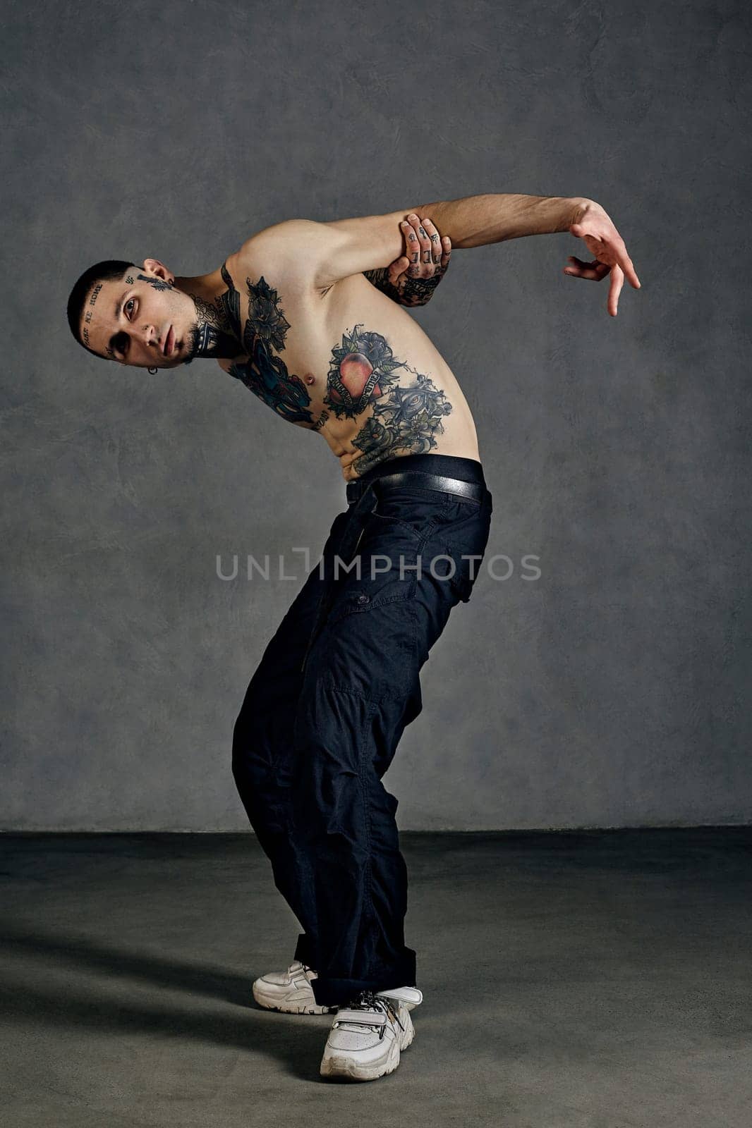 Athletic tattooed male with naked torso, beard. Dressed in black pants and white sneakers. Dancing against gray background. Dancehall, hip-hop by nazarovsergey