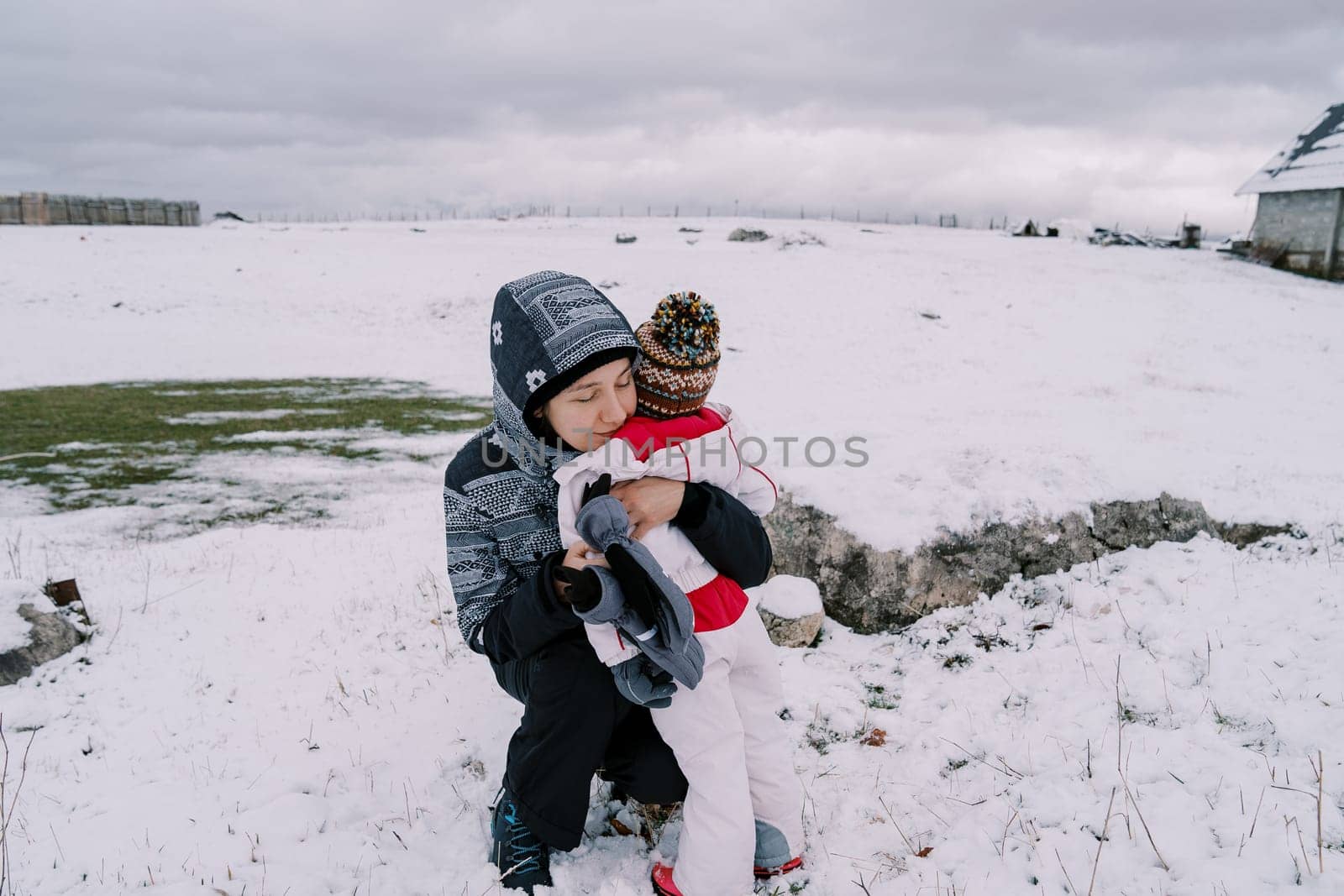 Mom hugs a little girl squatting on a snowy pasture near a farm by Nadtochiy